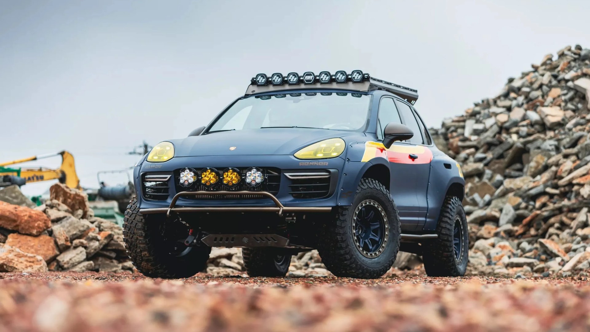 Matte blue off-road SUV with yellow headlights, roof lights, and rugged tires on rocky terrain.