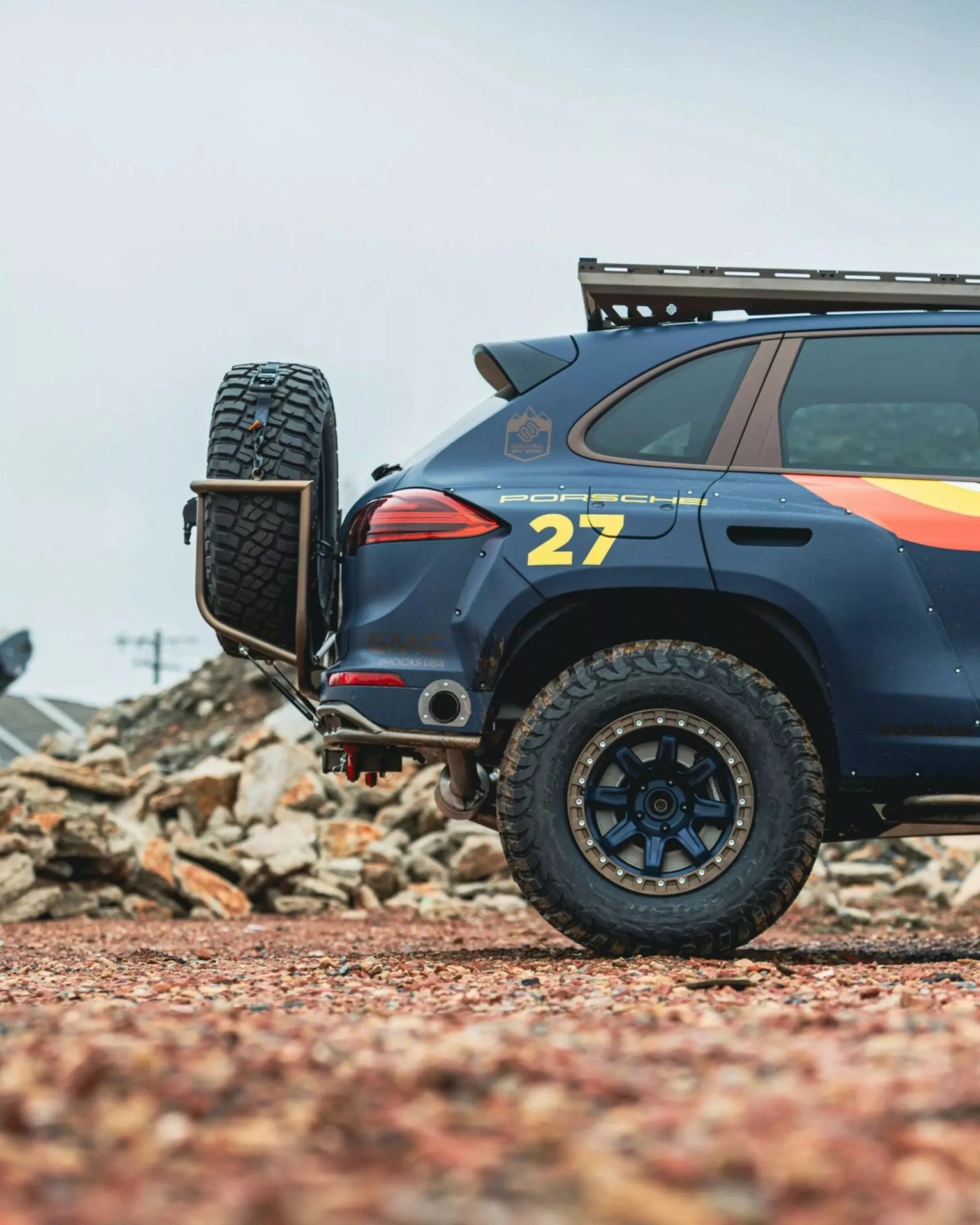 Rear side of a blue Porsche SUV with off-road tires, roof rack, spare tire mounted on the back, and yellow number 27 decal.