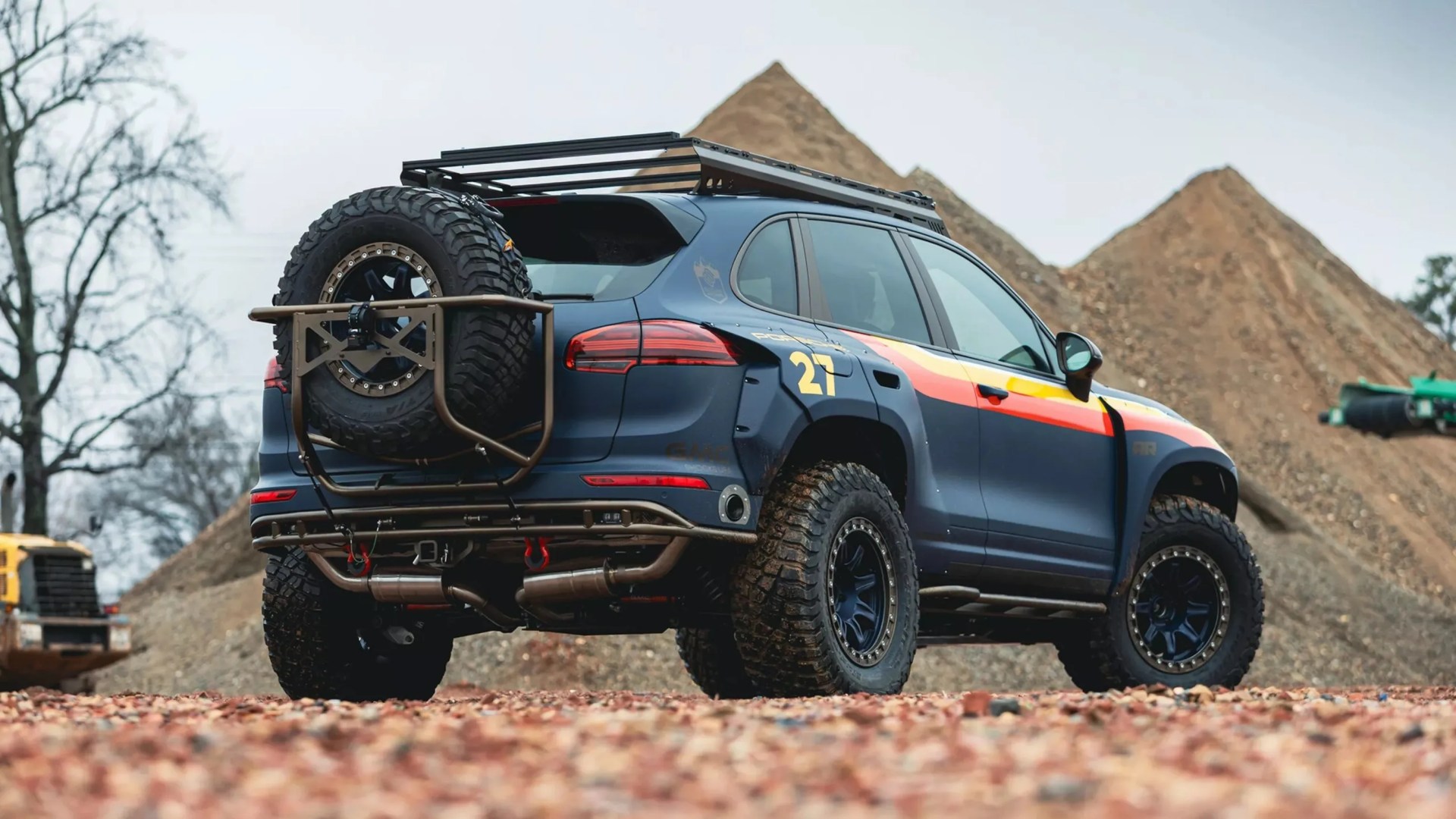 Matte blue off-road SUV with large knobby tires, roof rack, rear spare tire, and red-yellow stripe on the side.