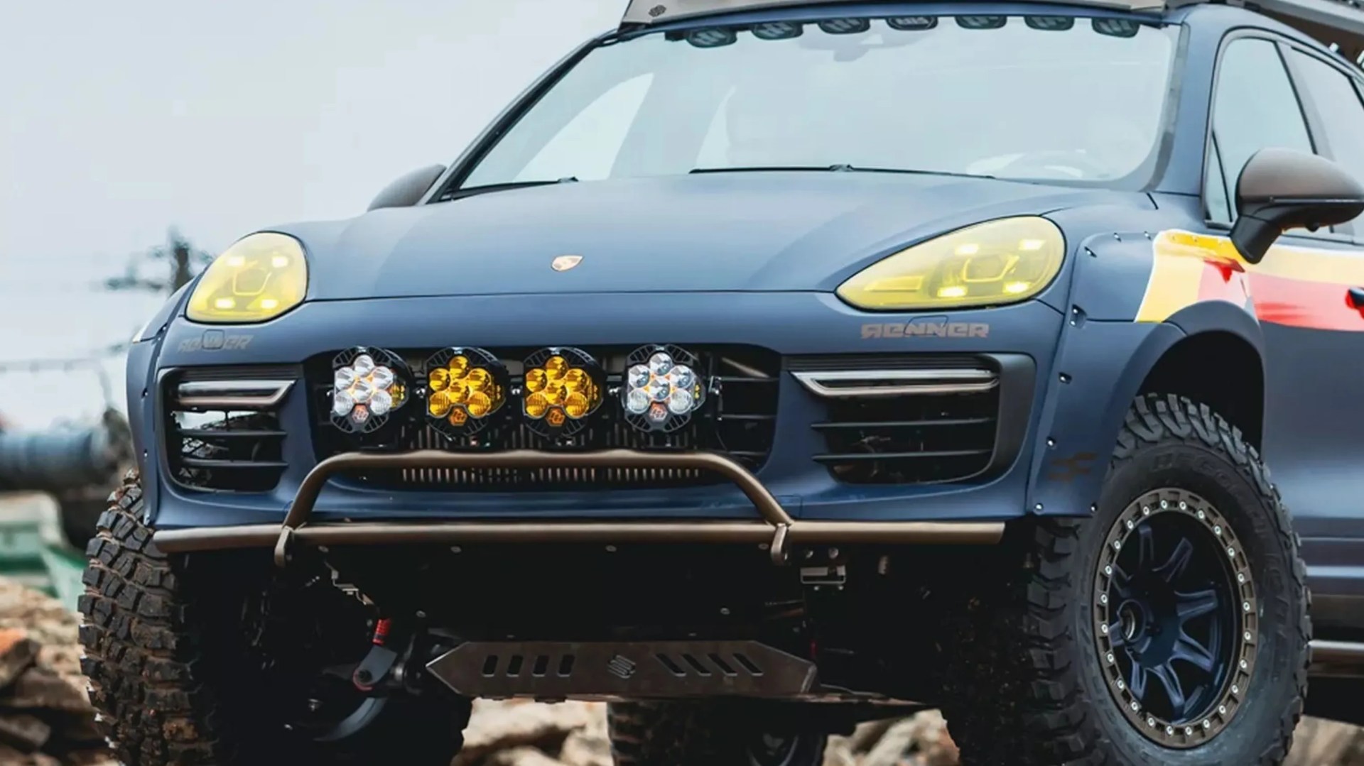 Blue Porsche SUV with off-road tires, yellow-tinted headlights, and four auxiliary lights mounted on the front grille.