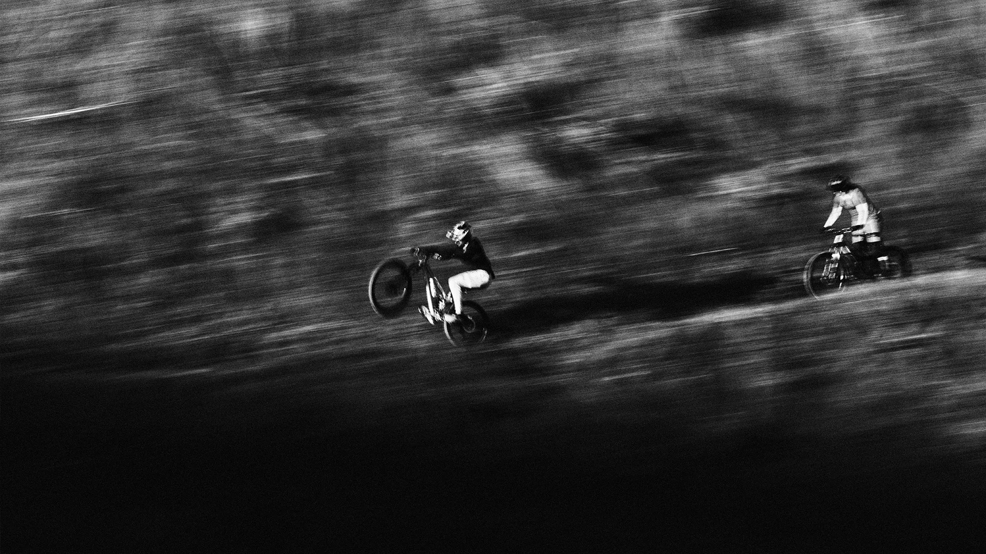 Two motorcyclists riding fast on a dirt trail, one performing a wheelie, in a blurred black and white photo.