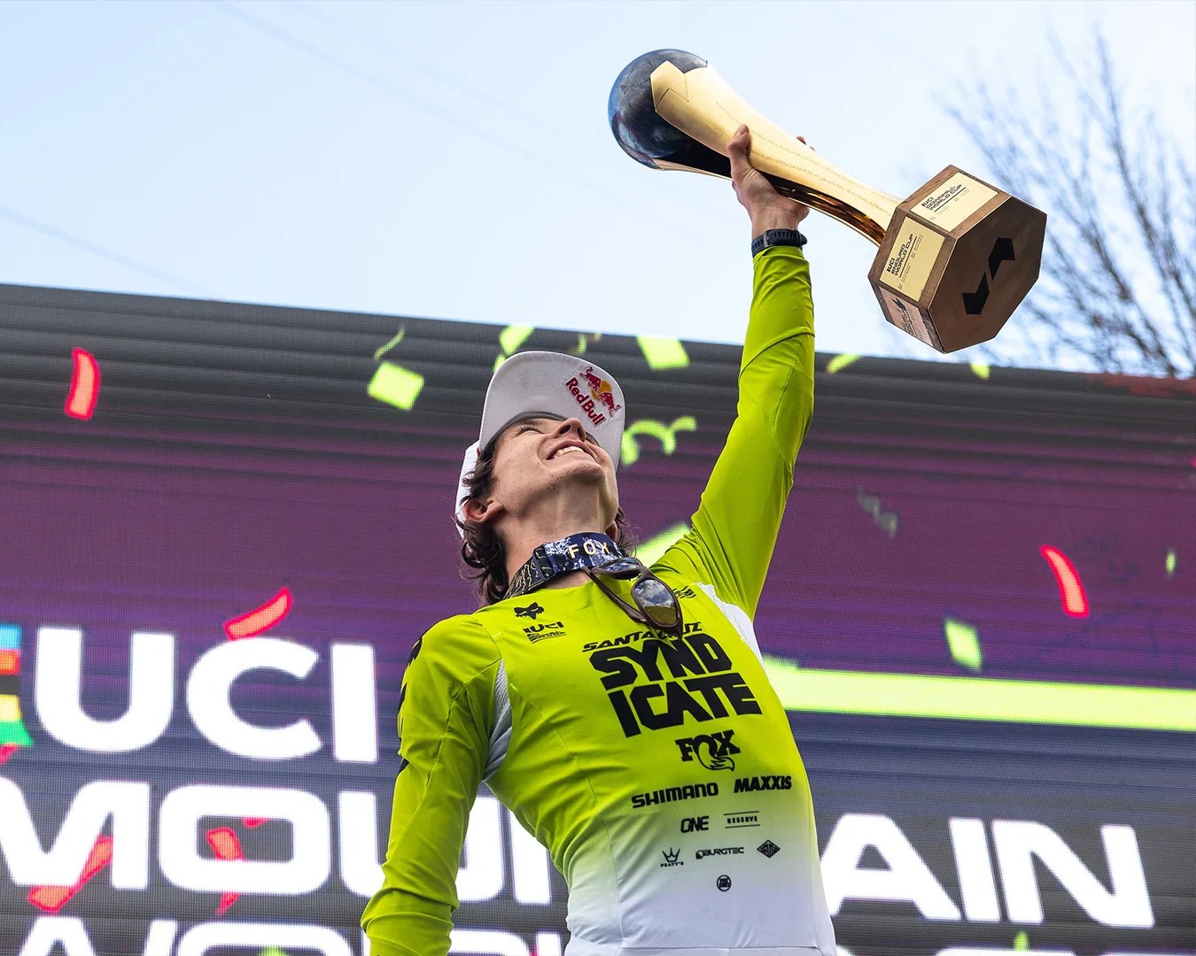 Person in a green and white cycling jersey holding a large gold trophy above their head with a Red Bull cap.