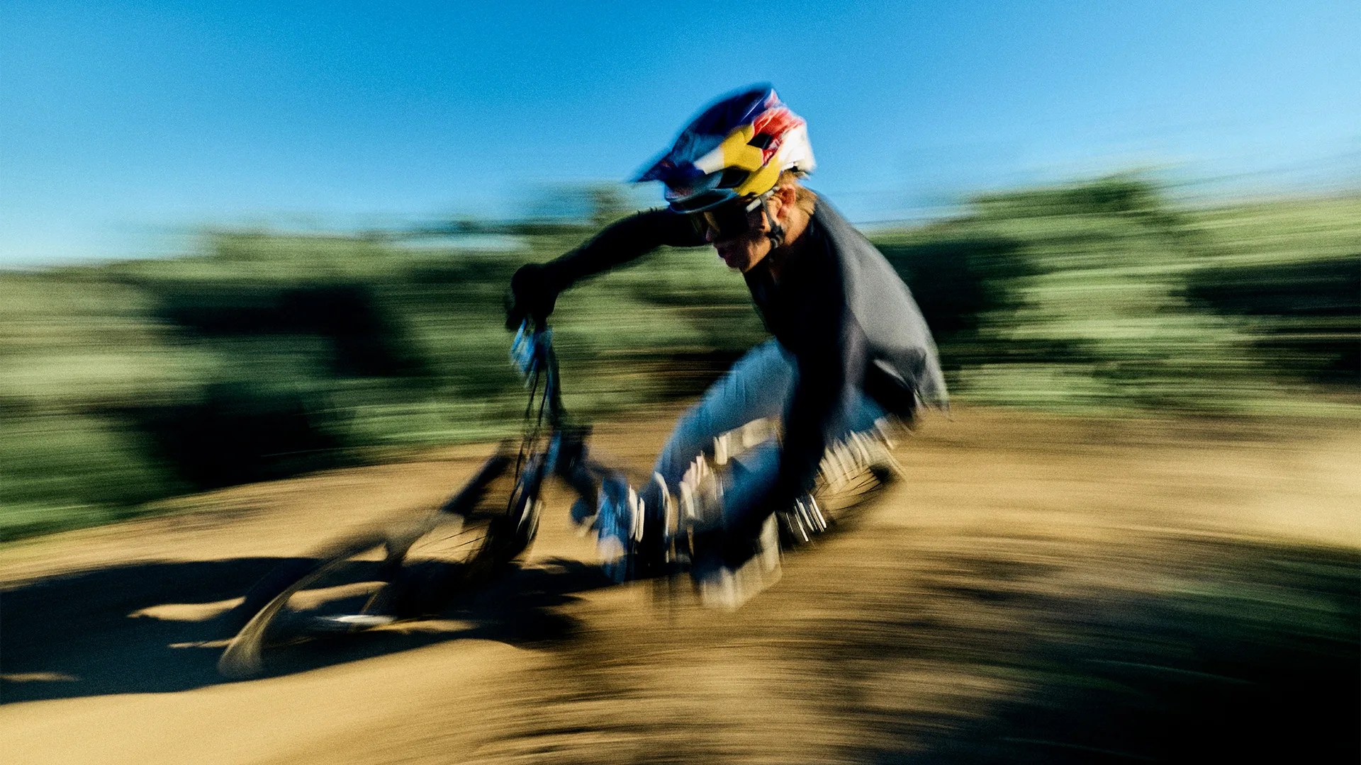 Mountain biker wearing a colorful helmet and black long sleeves riding fast on a dirt trail with motion blur.