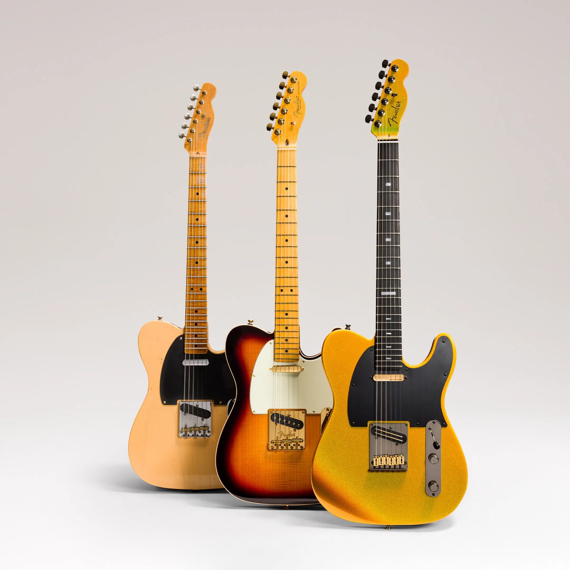 Three Fender Telecaster electric guitars in natural, sunburst, and gold sparkle finishes standing upright on a white surface.
