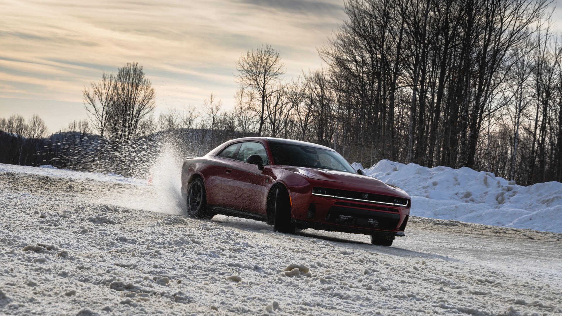 Red sports car drifting on a snowy road with snow spraying behind it and leafless trees in the background.
