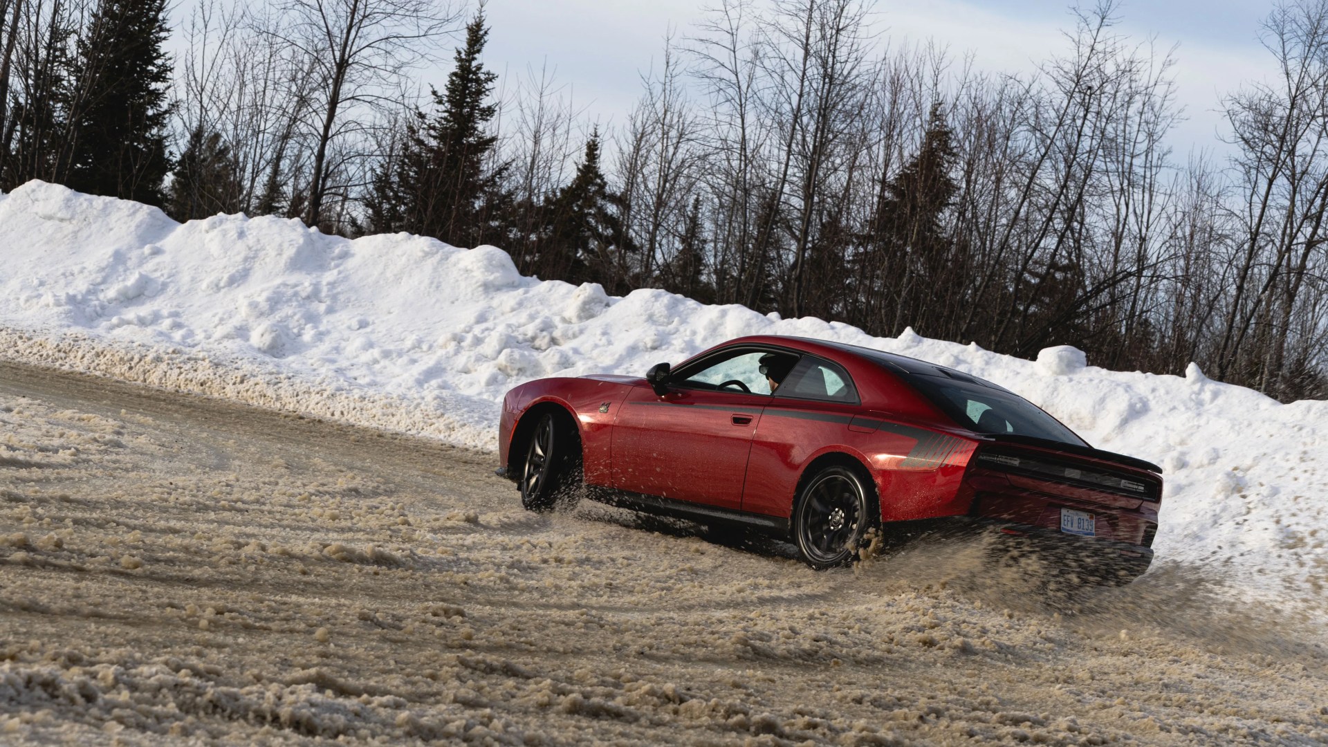 Red Dodge Challenger with black wheels driving on a snowy, muddy road with snowbanks and leafless trees in the background.