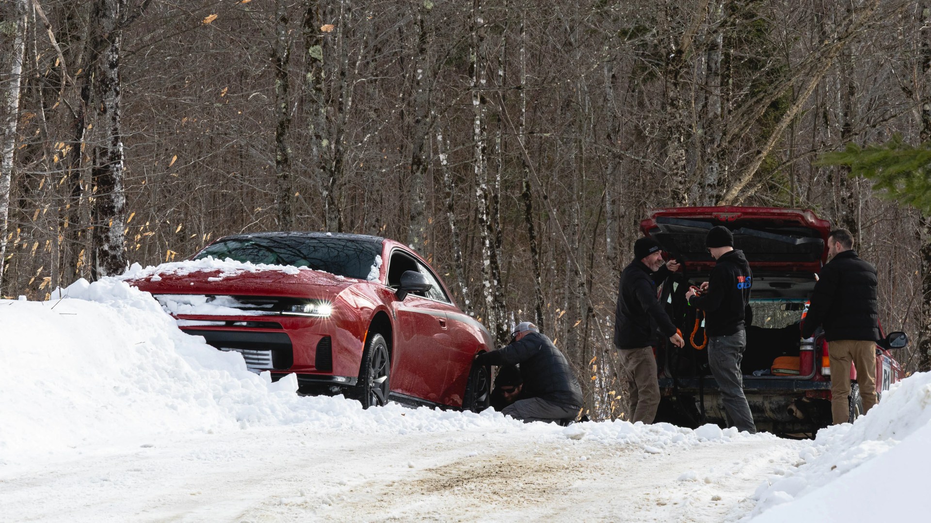 Red car stuck in snow with two people kneeling by the front tire and three people standing near an open trunk of a vehicle.