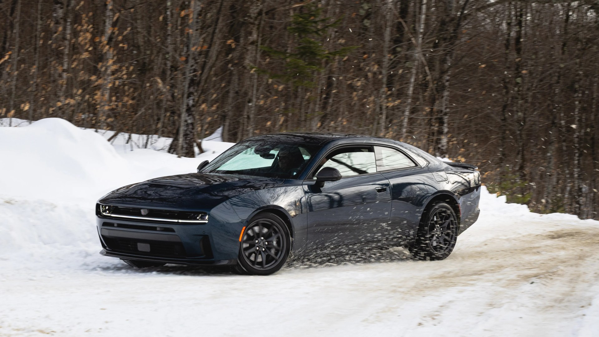 Dark blue two-door sports car driving on a snowy road with snow splashing from the rear tires.