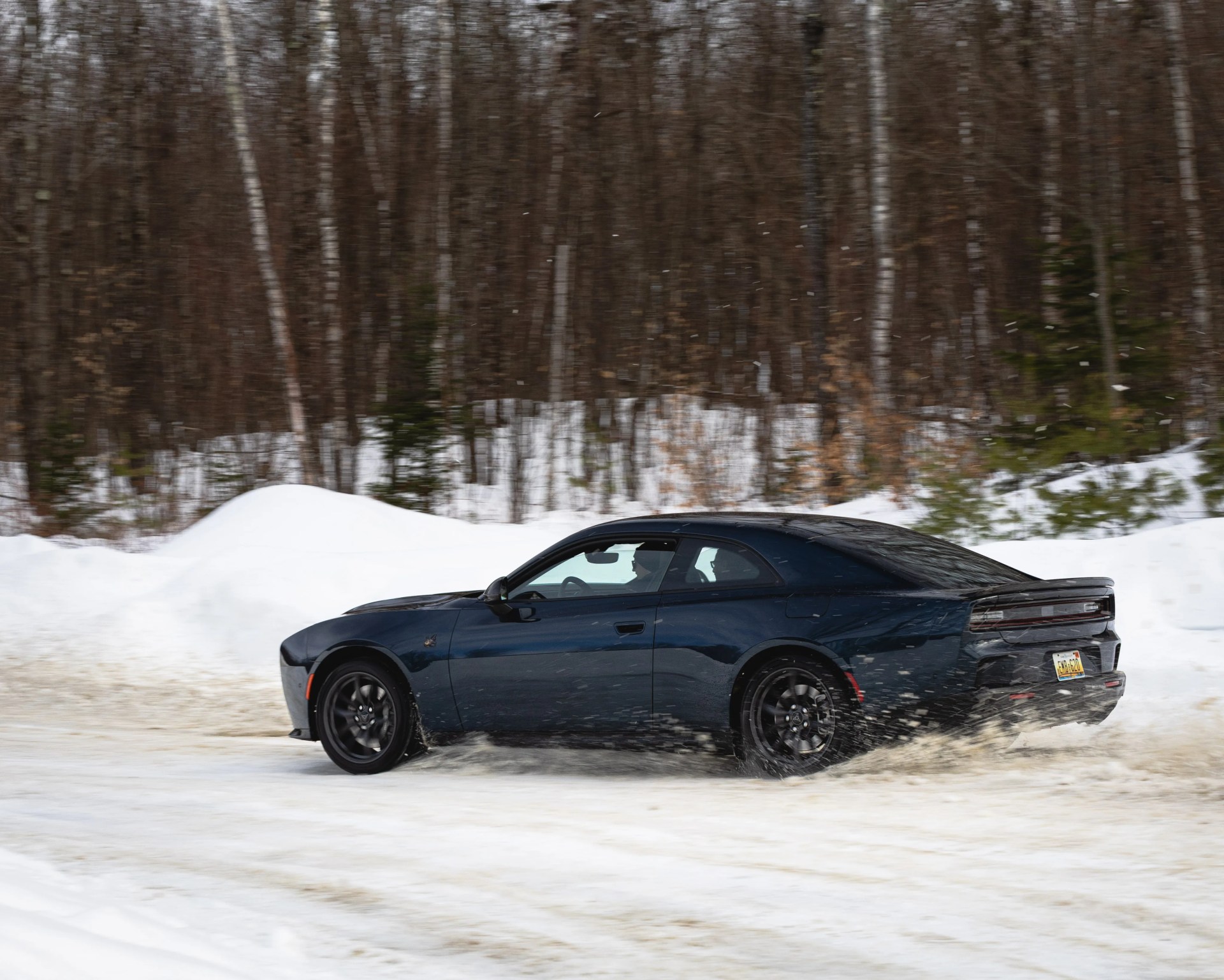 Black Dodge Charger driving on a snowy road with snow-covered trees in the background.