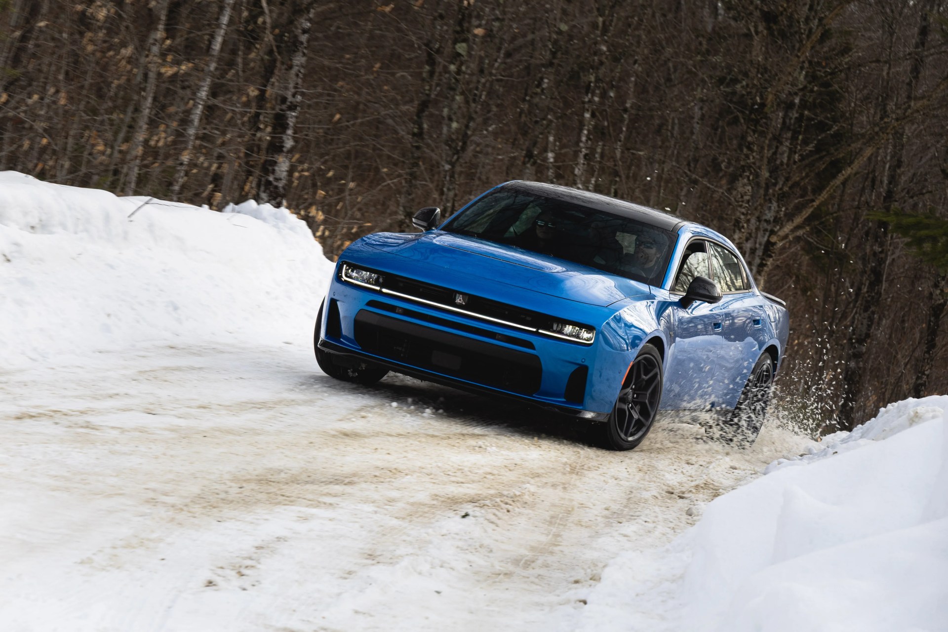 Blue sedan with black roof drifting on a snowy road with snowbanks and bare trees in the background.