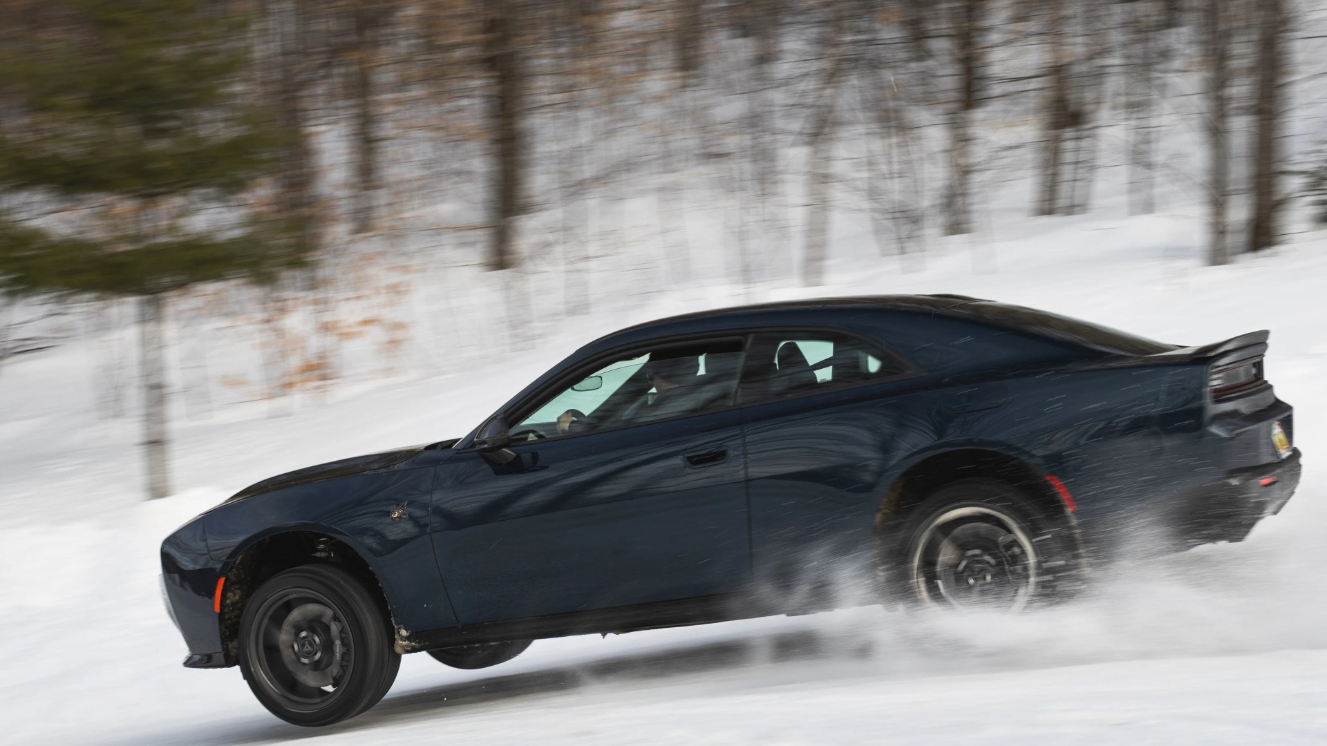 Dark blue Dodge Charger with black wheels drifting on snow-covered ground with blurred trees in the background.