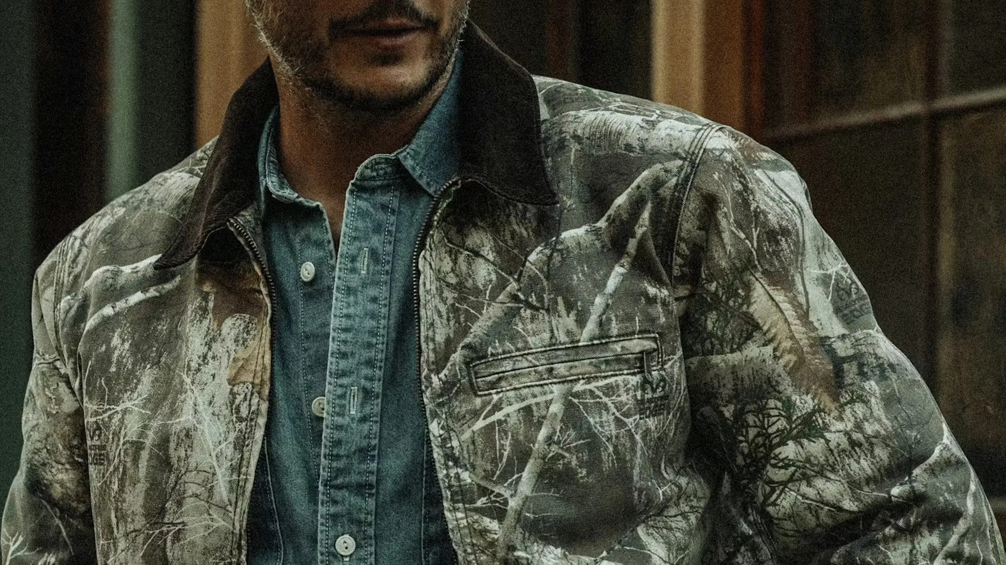 Man wearing a camouflage jacket with a dark collar over a blue denim button-up shirt.