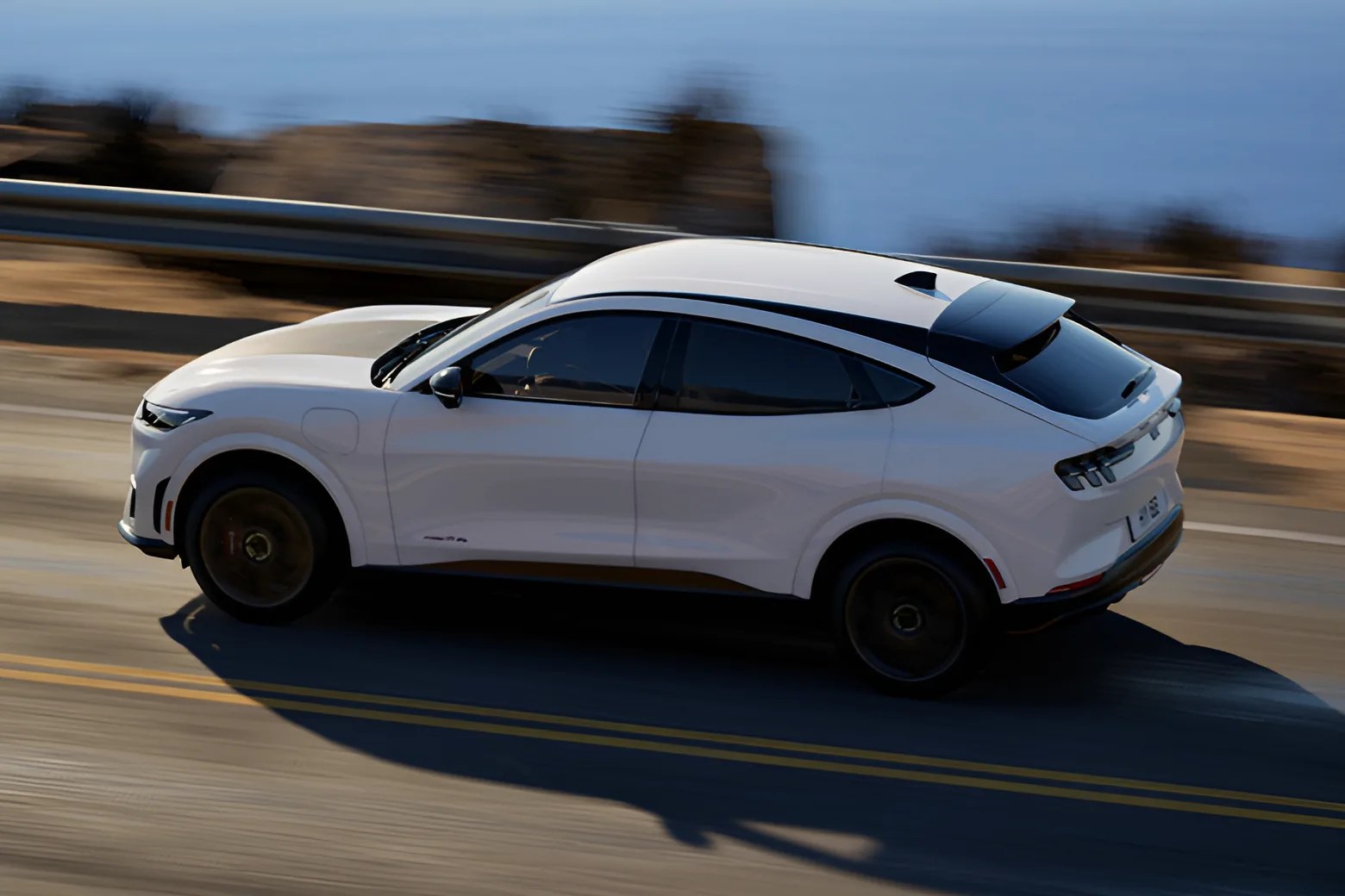 White electric SUV driving on a coastal road with tinted windows and black roof spoiler.