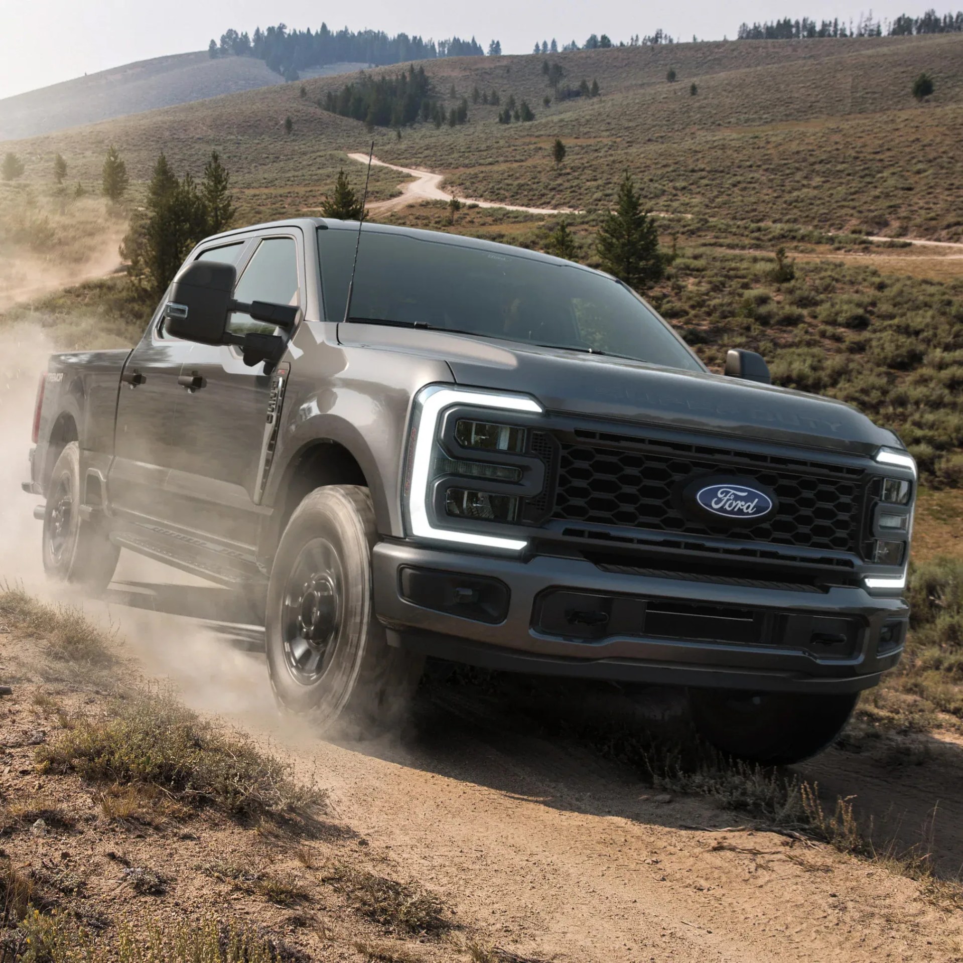 Gray Ford pickup truck driving on a dusty dirt road in a hilly, grassy landscape.