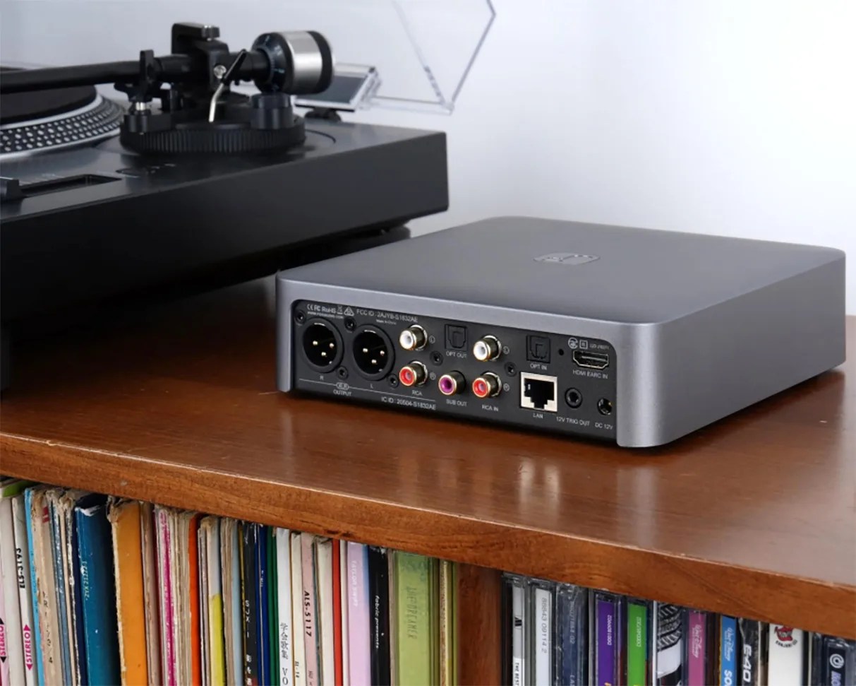 Gray audio interface with multiple input/output ports on a wooden shelf next to a black turntable.
