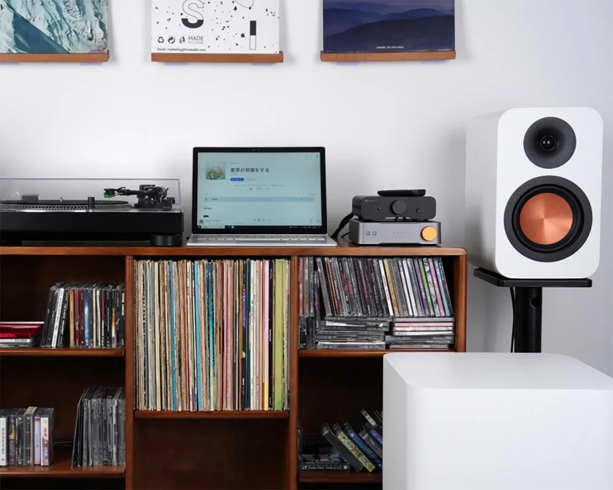 White bookshelf speaker with copper driver on a black stand next to a wooden shelf holding vinyl records, a laptop, and audio equipment.