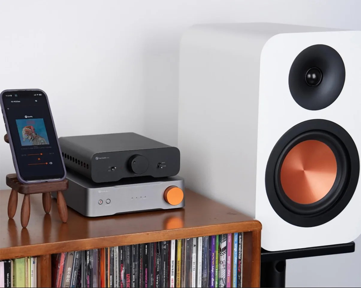 White speaker with copper cone driver next to black and silver audio equipment with orange knob on wooden shelf with CDs.