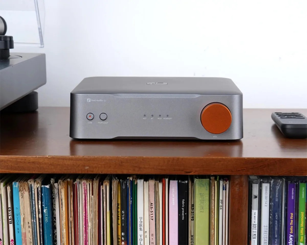 Compact silver audio amplifier with an orange volume knob on a wooden shelf above a collection of vinyl records.