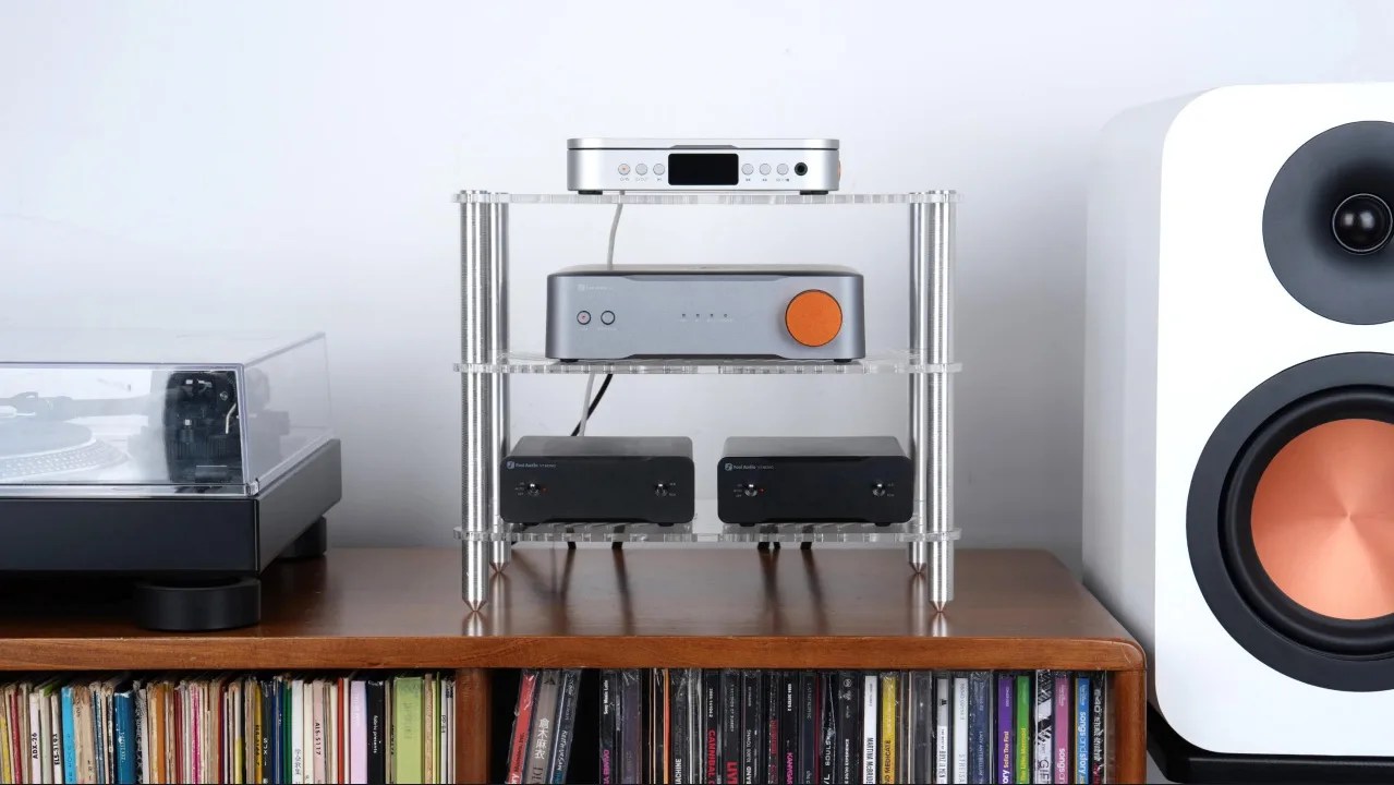 Audio equipment including a turntable, amplifier with orange dial, two black audio units on a clear acrylic stand, and a white speaker with copper cone on a wooden shelf.
