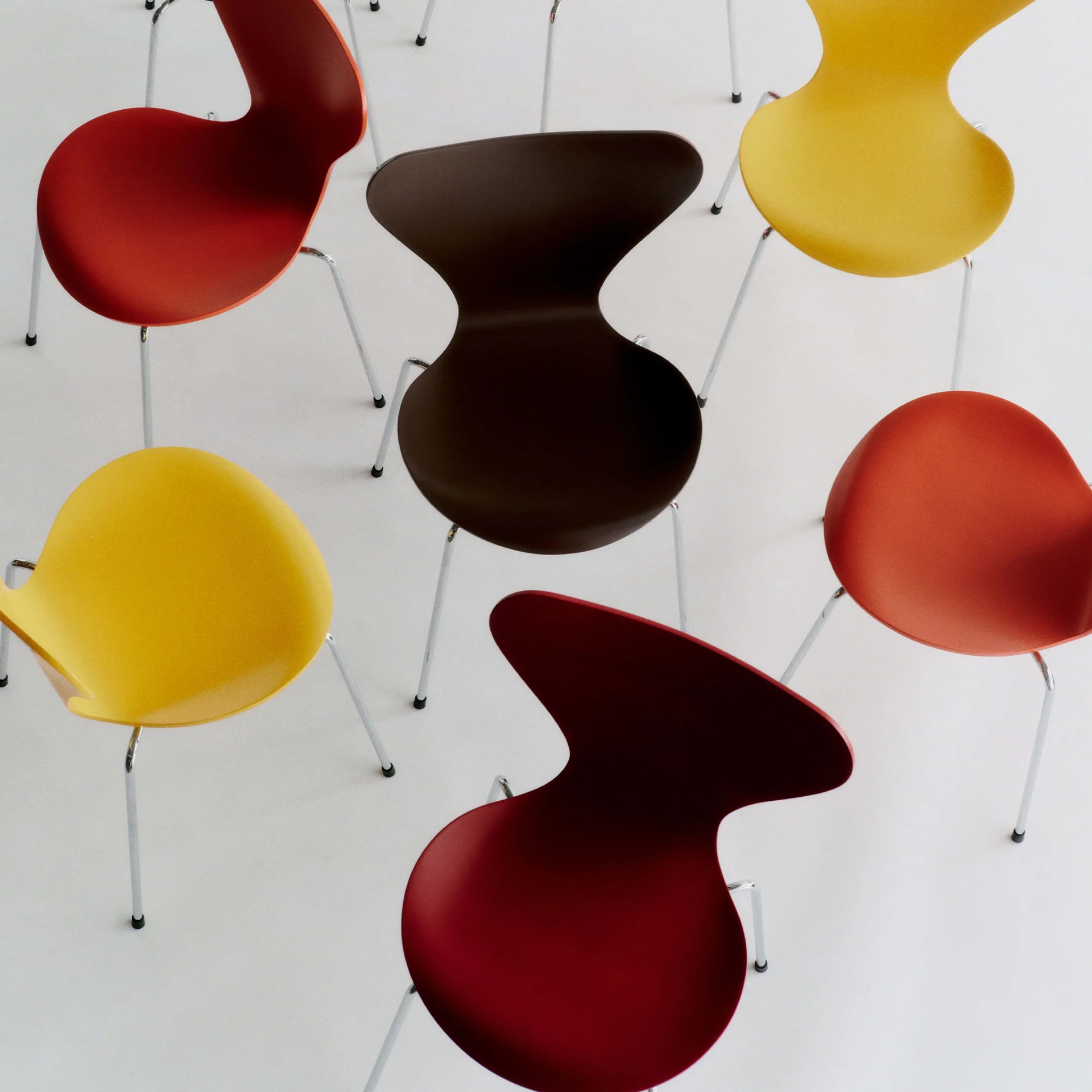 Six modern chairs with curved seats in red, yellow, and dark brown, each with metal legs on a white floor.