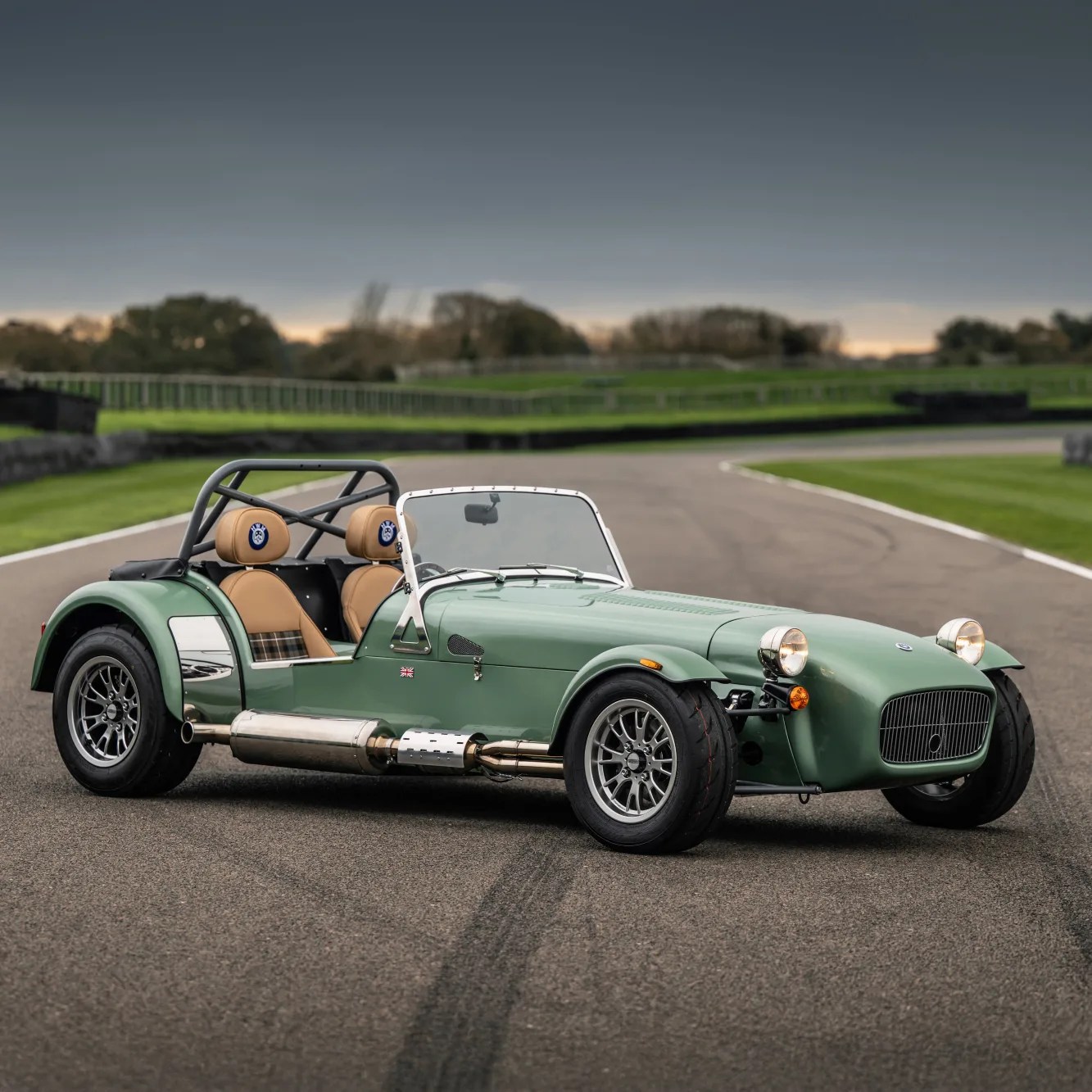 Green vintage-style open-top sports car with tan leather seats and chrome exhaust on a racetrack.