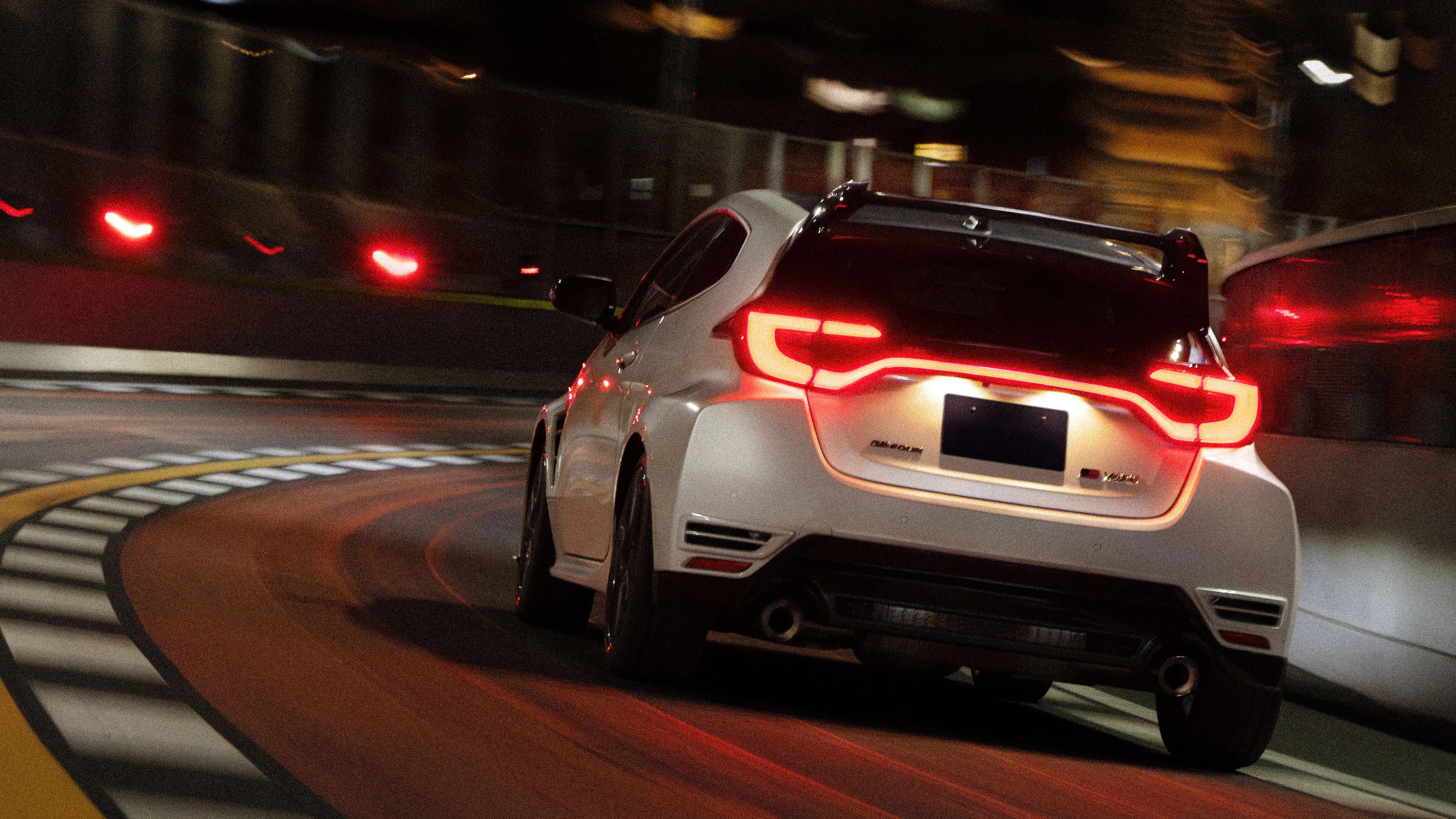 White sporty hatchback car with illuminated taillights driving on a curved racetrack at night.