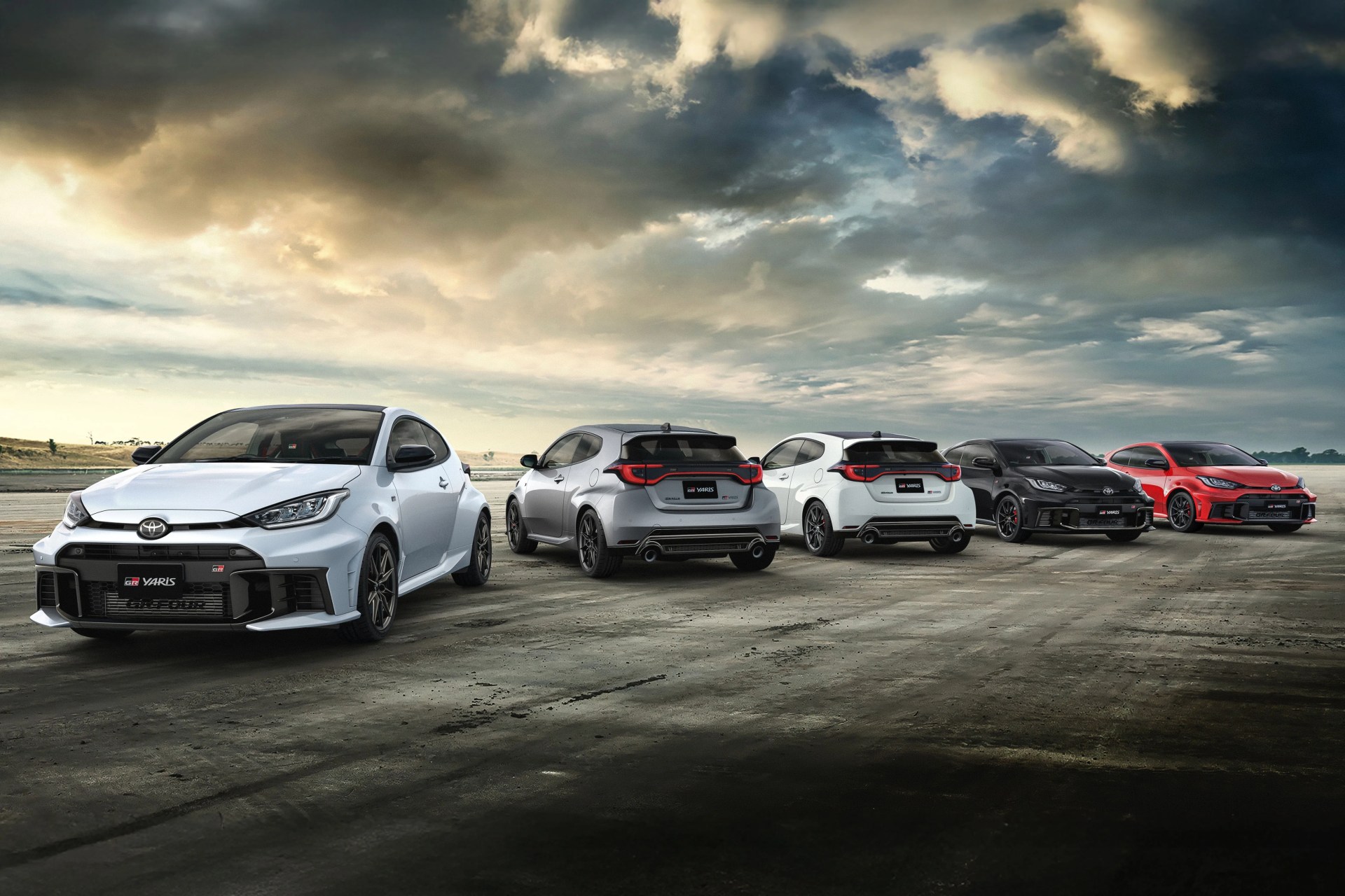 Five Toyota GR Yaris cars in white, silver, black, and red parked on a wide open concrete surface under a cloudy sky.