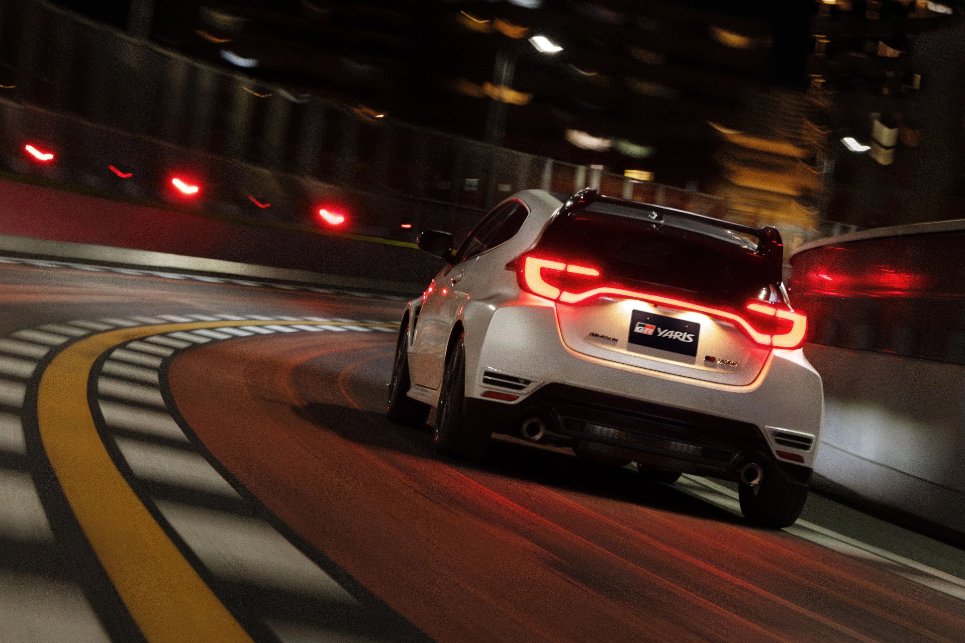 White Toyota GR Yaris hatchback with illuminated taillights driving on a curved racetrack at night.