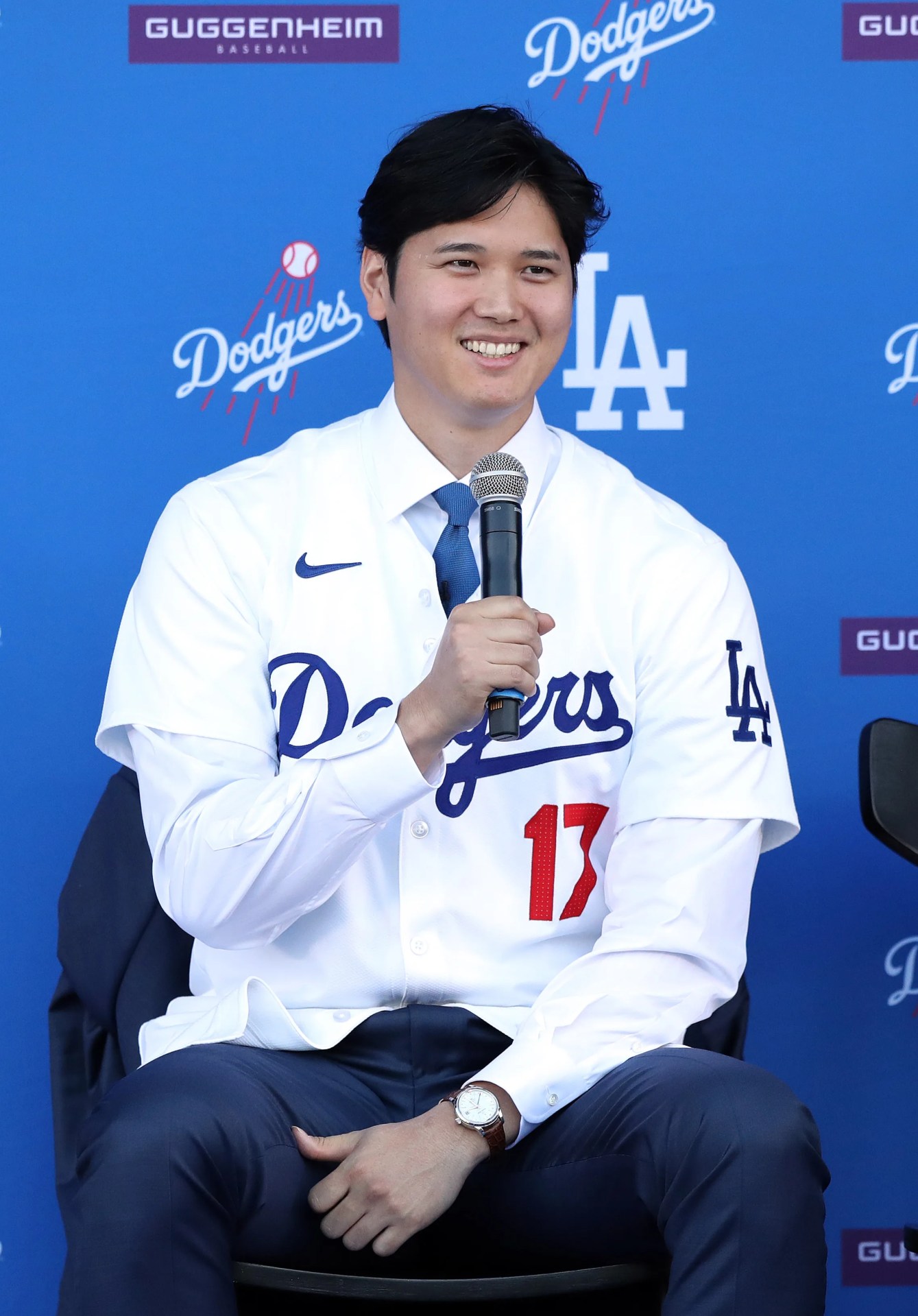 Man wearing a white Dodgers baseball jersey with number 17, blue tie, and holding a microphone while seated.