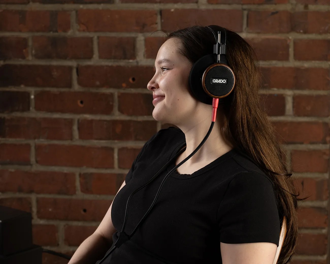Person wearing black Grado over-ear headphones with wooden accents against a brick wall background.