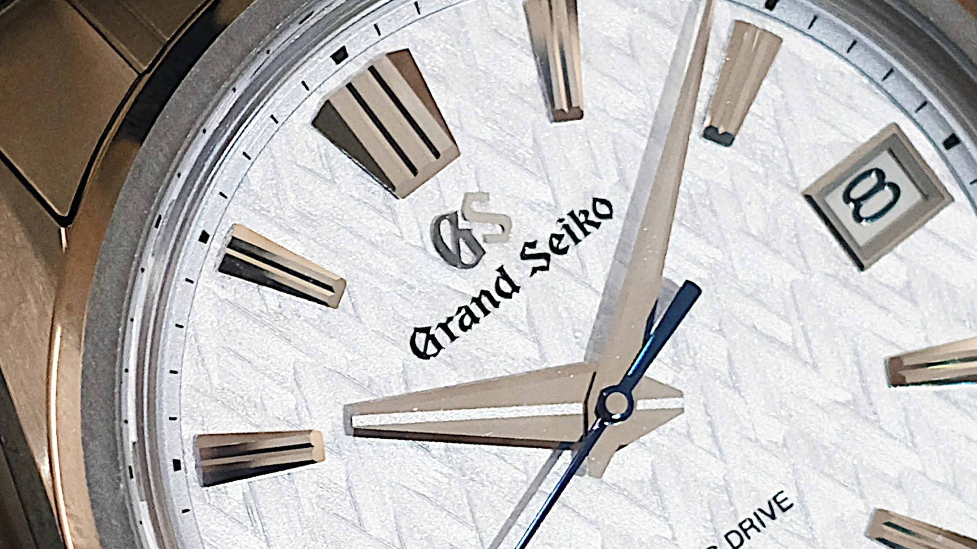 Close-up of a Grand Seiko watch face with silver hour markers, date window at 3 o'clock, and textured white dial.