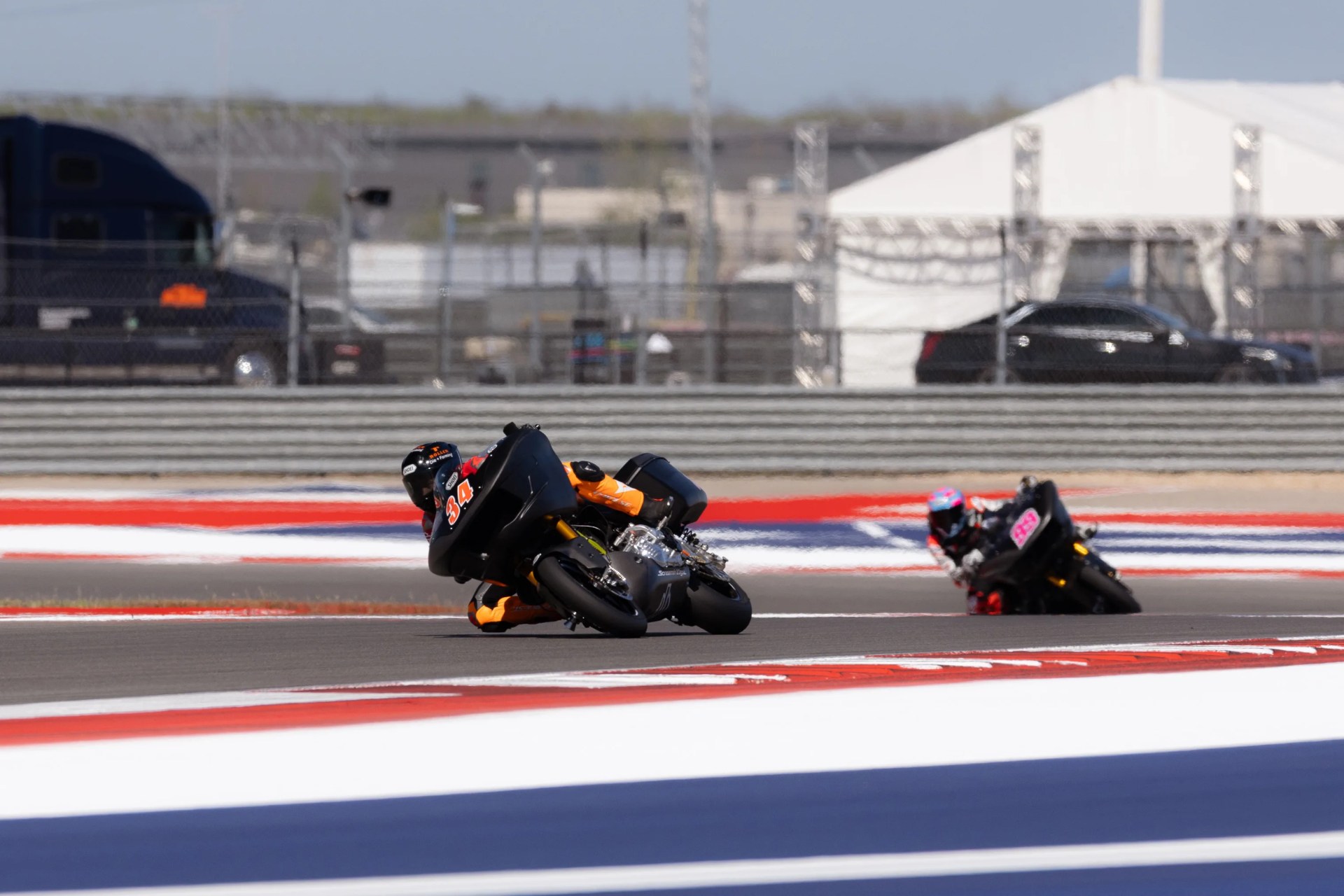 Two motorcyclists leaning sharply on black racing bikes on a track with red, white, and blue markings.