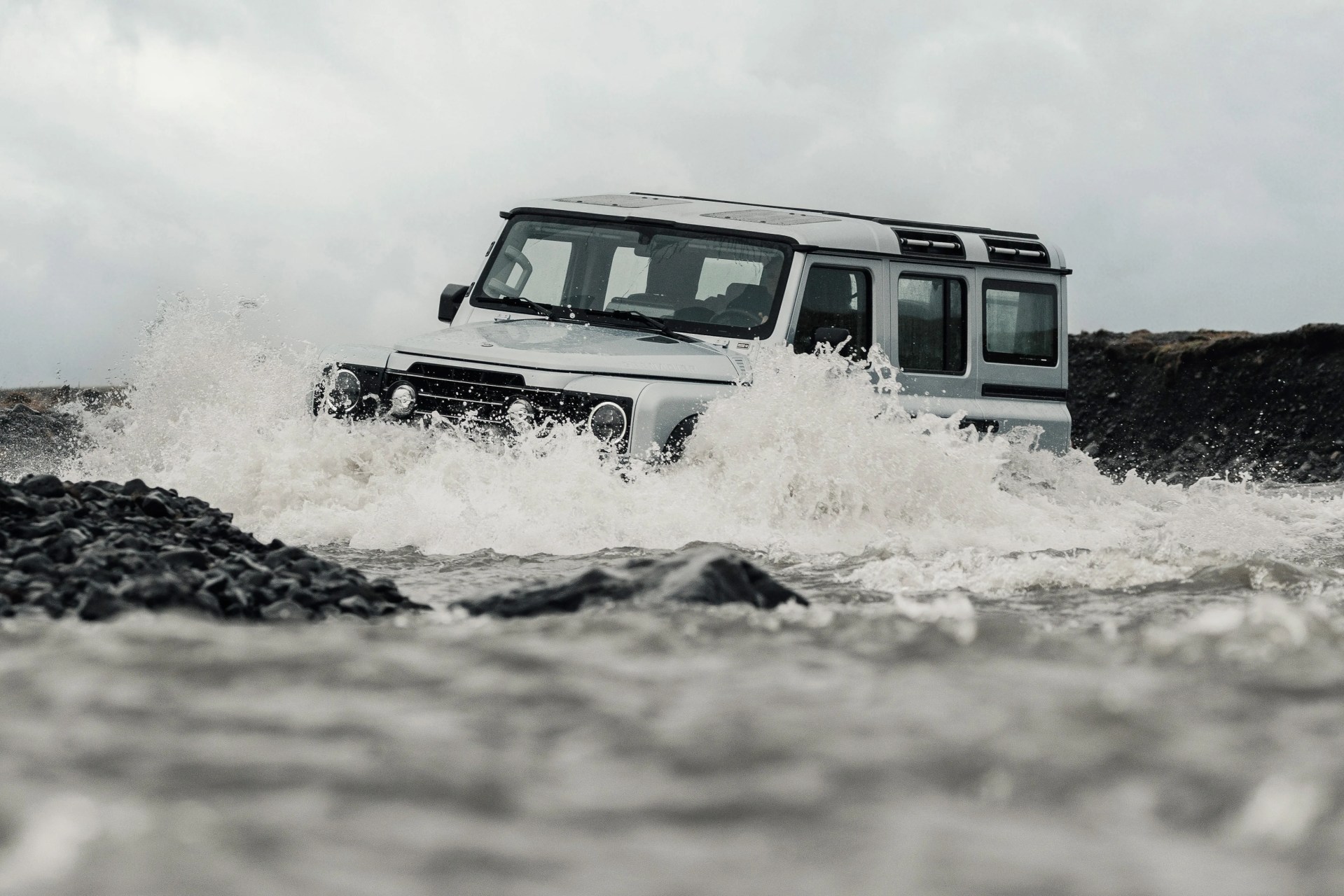 Silver off-road SUV driving through a rocky river with water splashing around it.