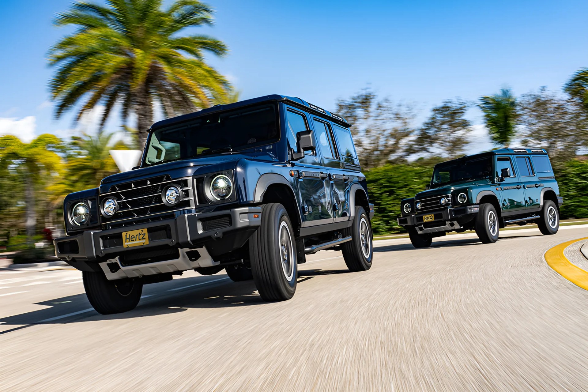 Two dark blue Land Rover Defenders driving on a curved road with palm trees and greenery in the background.
