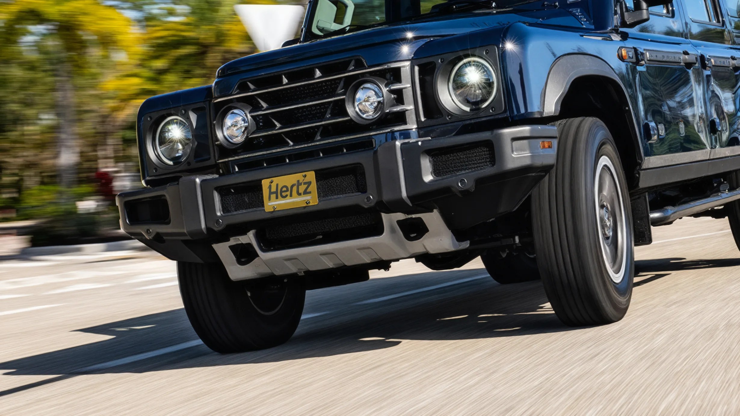 Front view of a black off-road vehicle with a yellow Hertz license plate driving on a road.