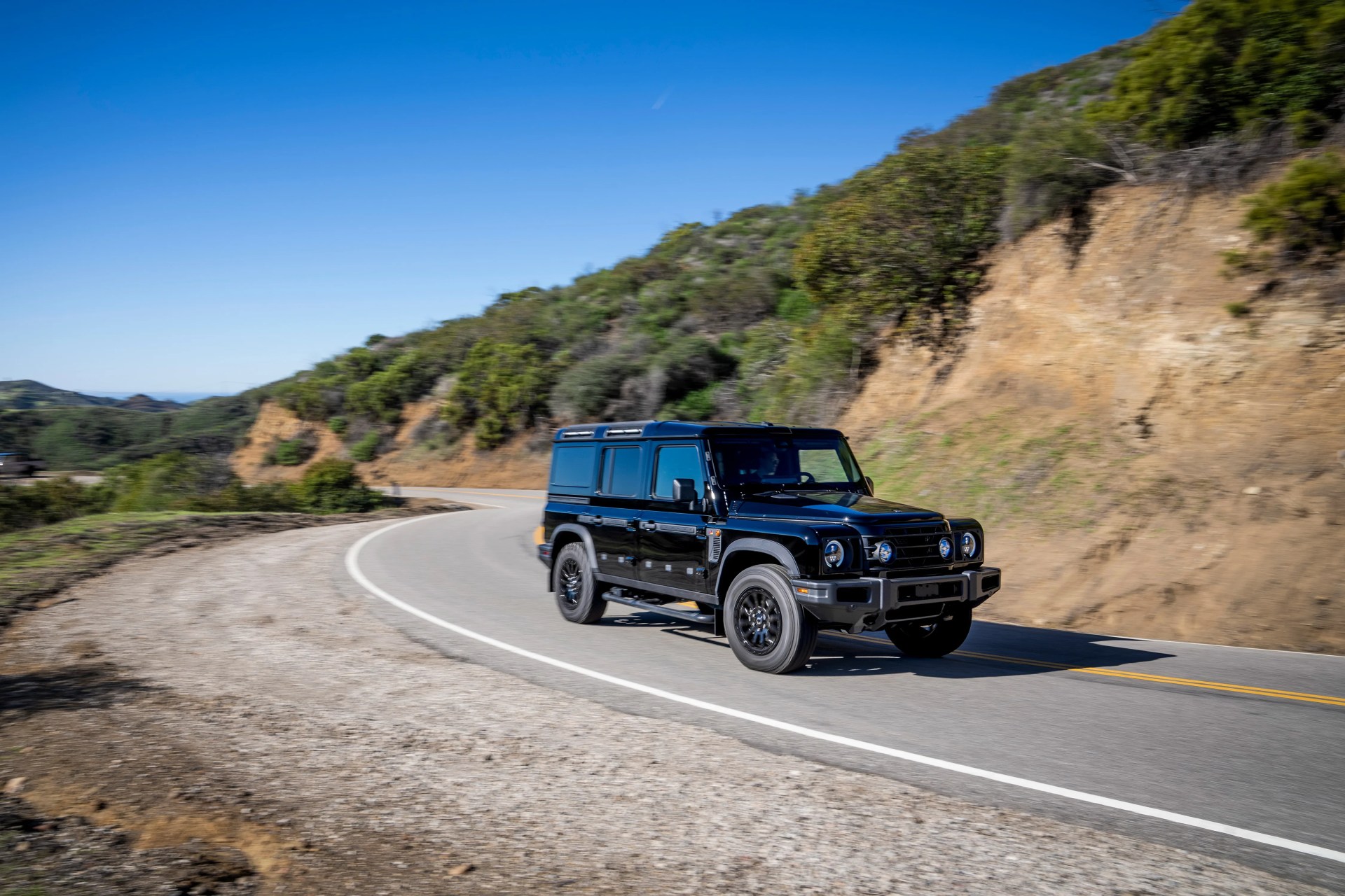 Black off-road SUV driving on a winding mountain road with dry vegetation and clear blue sky.