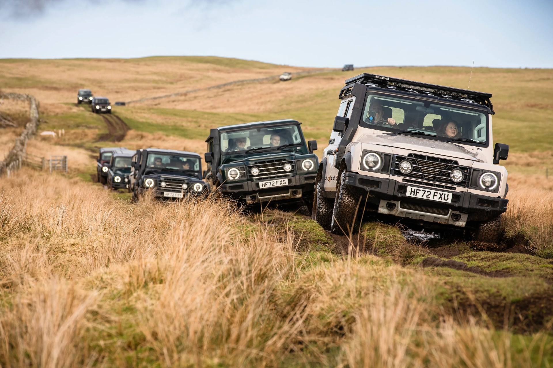 Line of off-road SUVs driving on a muddy grassy trail in a hilly countryside.
