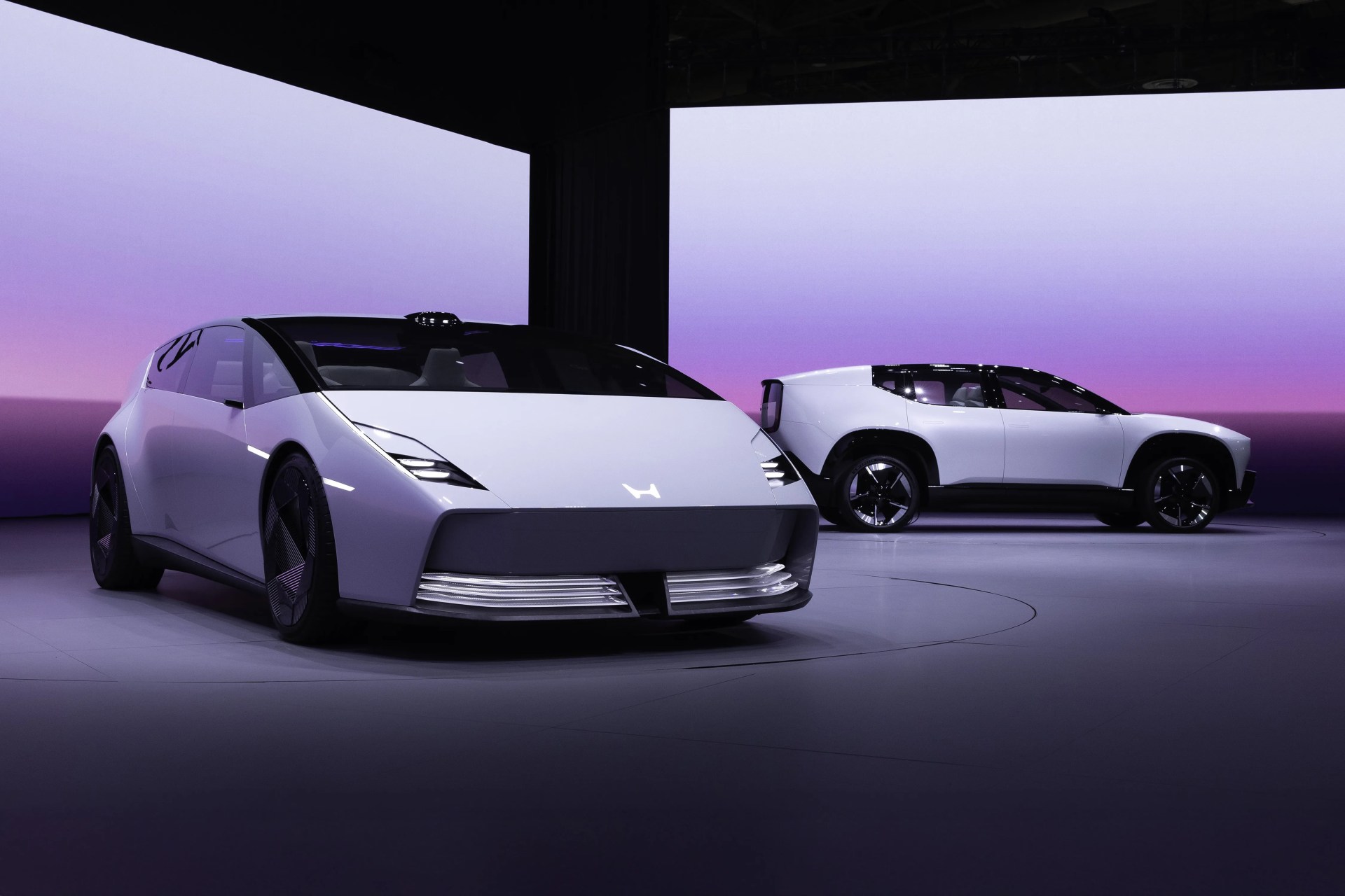 Two white futuristic electric cars with sleek designs displayed indoors against a purple gradient backdrop.