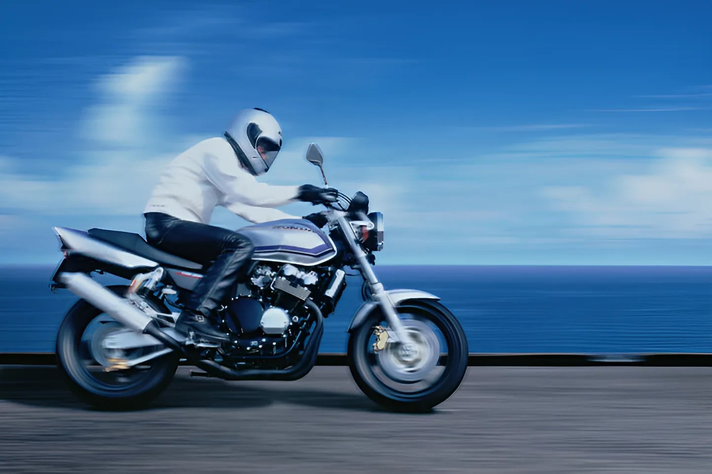 Person in white helmet and jacket riding a silver motorcycle on a road by the ocean.