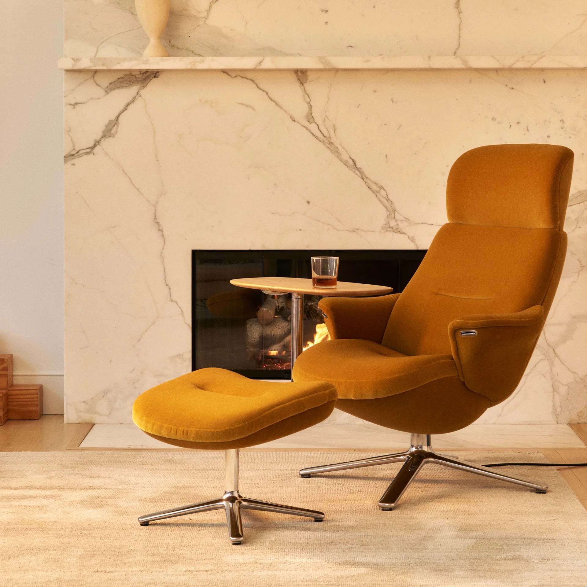 Mustard yellow upholstered lounge chair with matching ottoman and attached wooden side table, set in front of a marble fireplace.