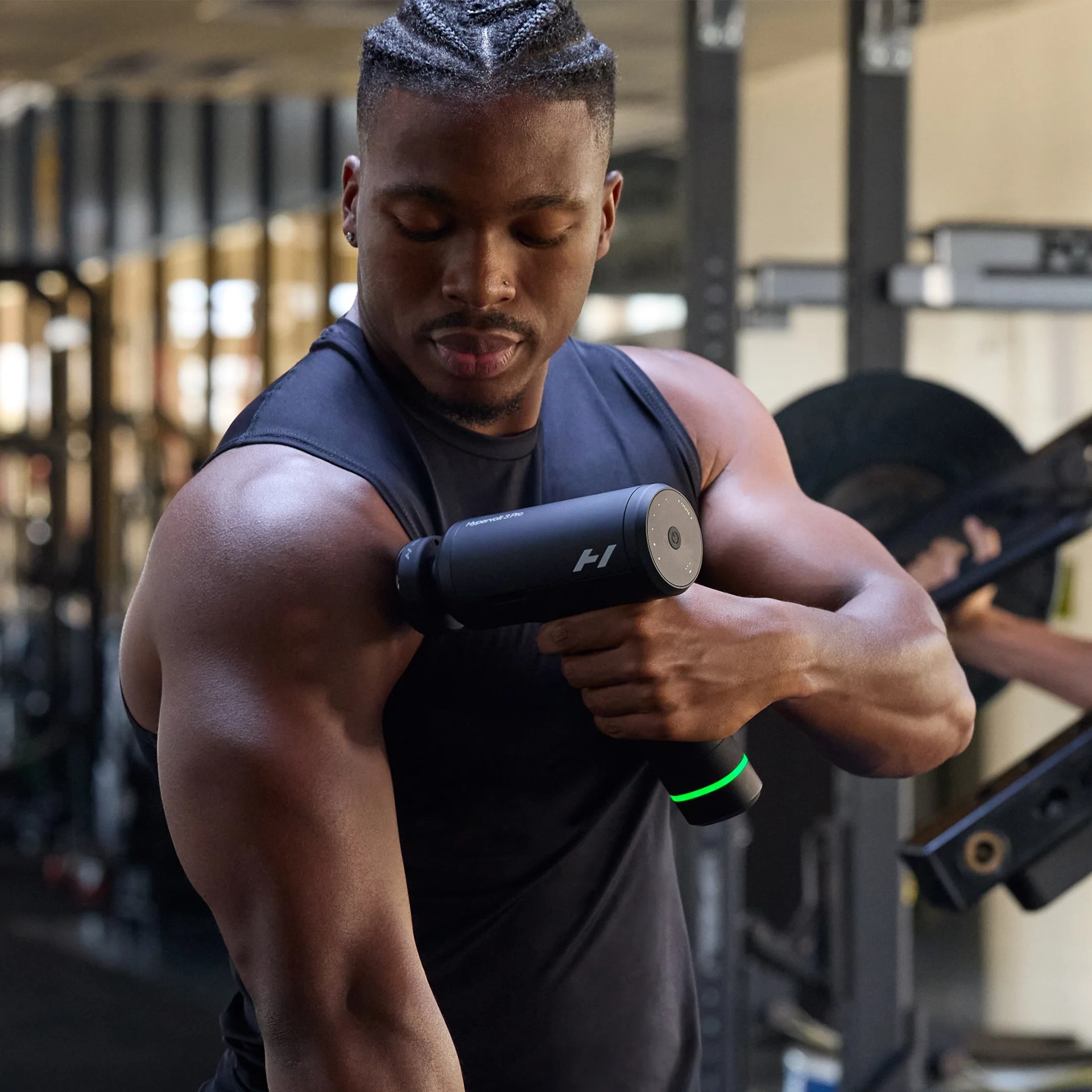 Man in a black sleeveless shirt using a black handheld massage gun on his shoulder in a gym.