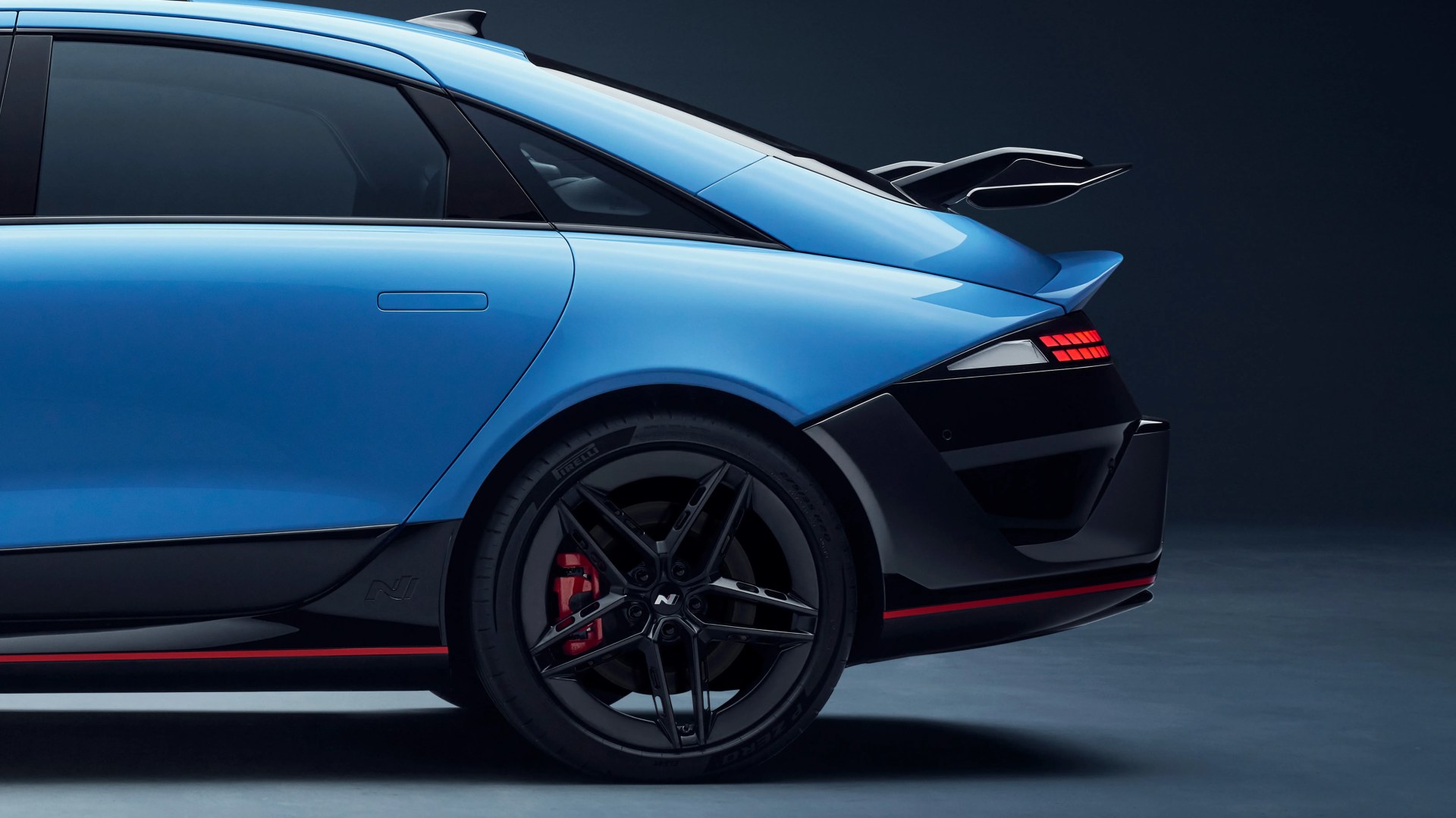 Rear side view of a blue sports car with black and red accents, black alloy wheel, and raised rear spoiler.