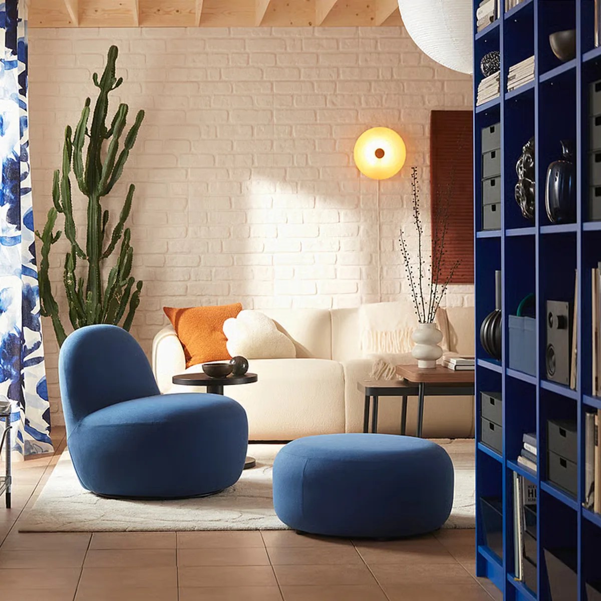 Blue upholstered rounded lounge chair and matching ottoman on a light rug in a living room with a white sofa and tall cactus.