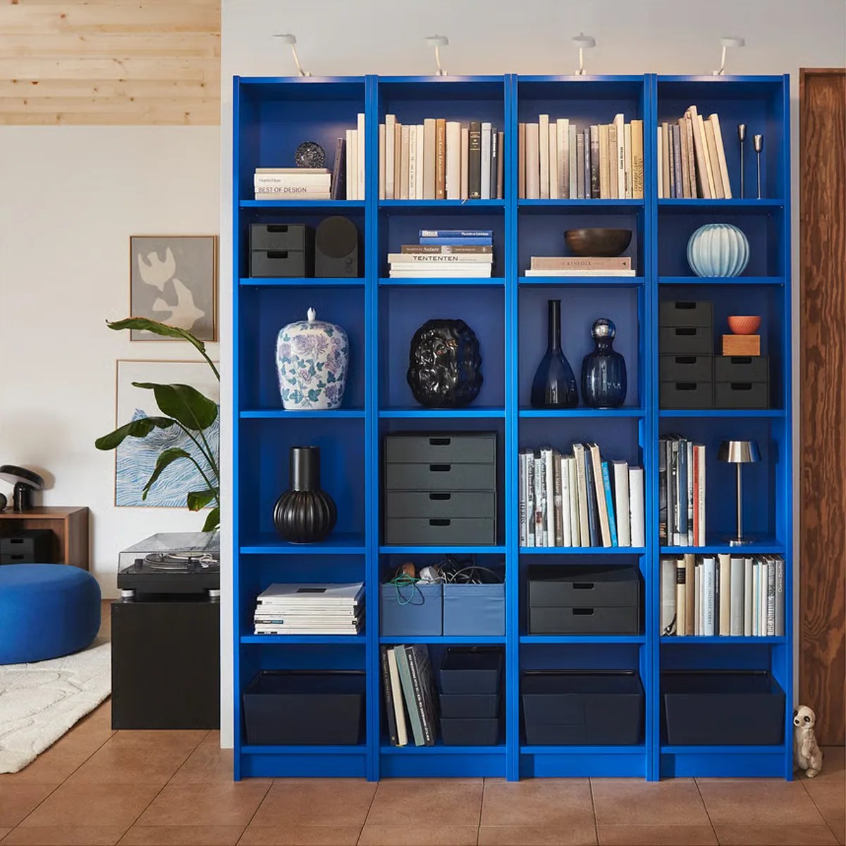 Tall blue bookshelf with multiple compartments holding books, black storage boxes, decorative vases, and small lamps.