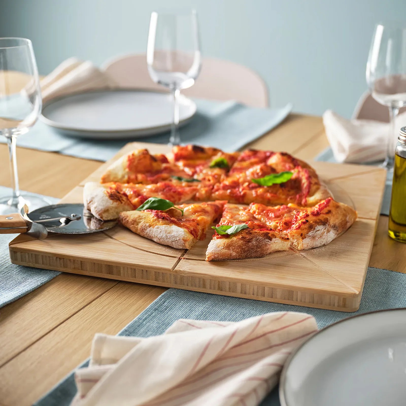 Wooden pizza board with a sliced margherita pizza topped with basil leaves on a dining table.