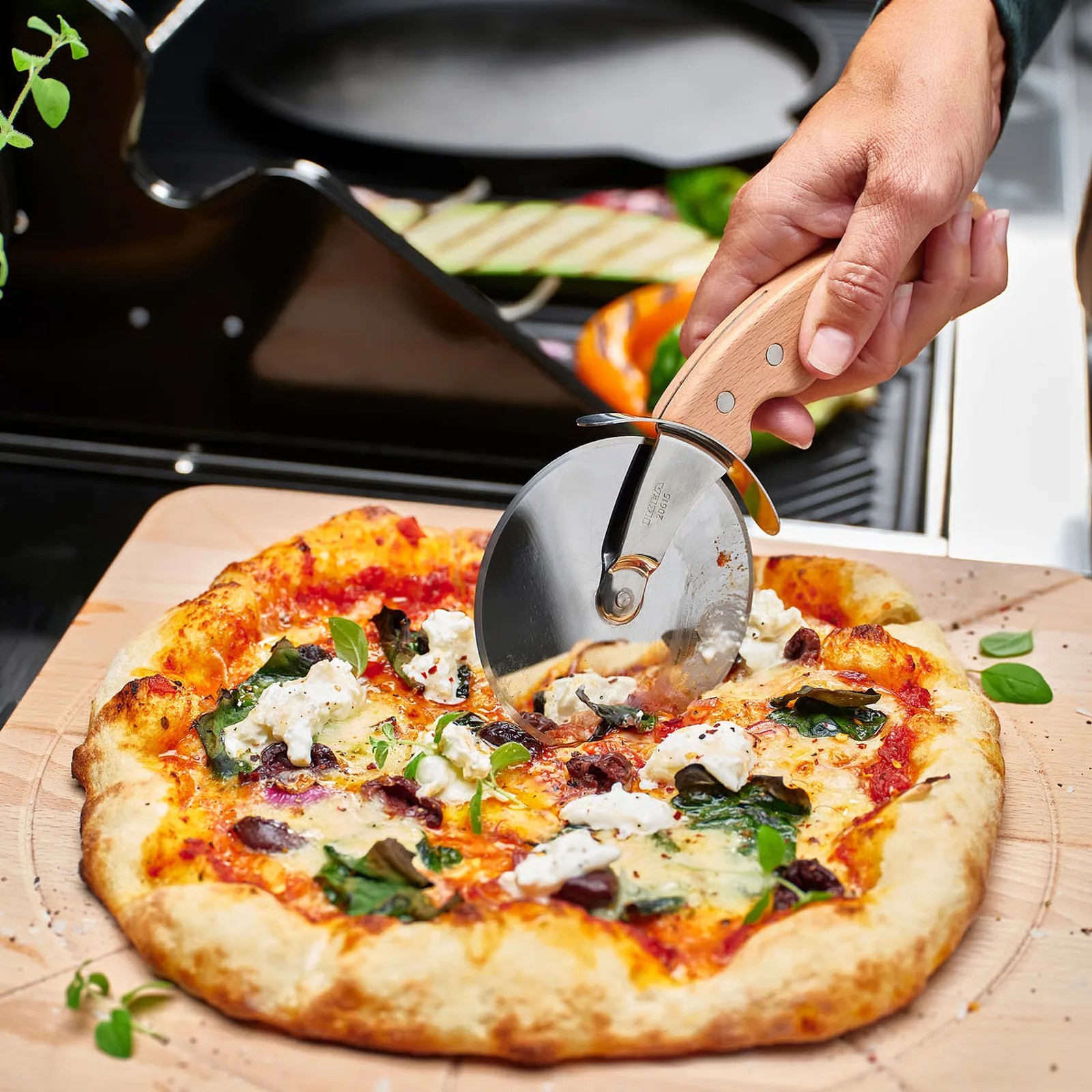 Hand holding a wooden-handled pizza cutter slicing a pizza with cheese, spinach, and olives on a wooden board.