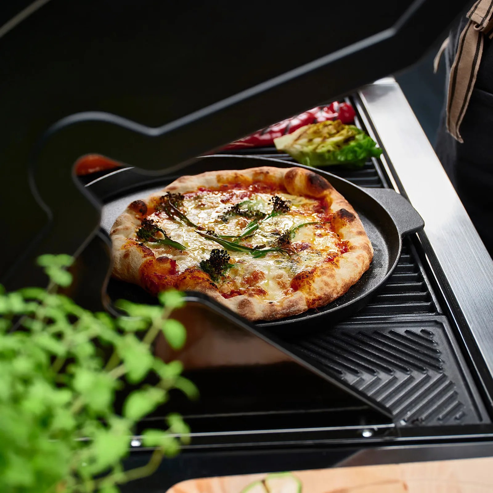 Cast iron pizza pan with freshly baked pizza topped with cheese and greens on a grill.