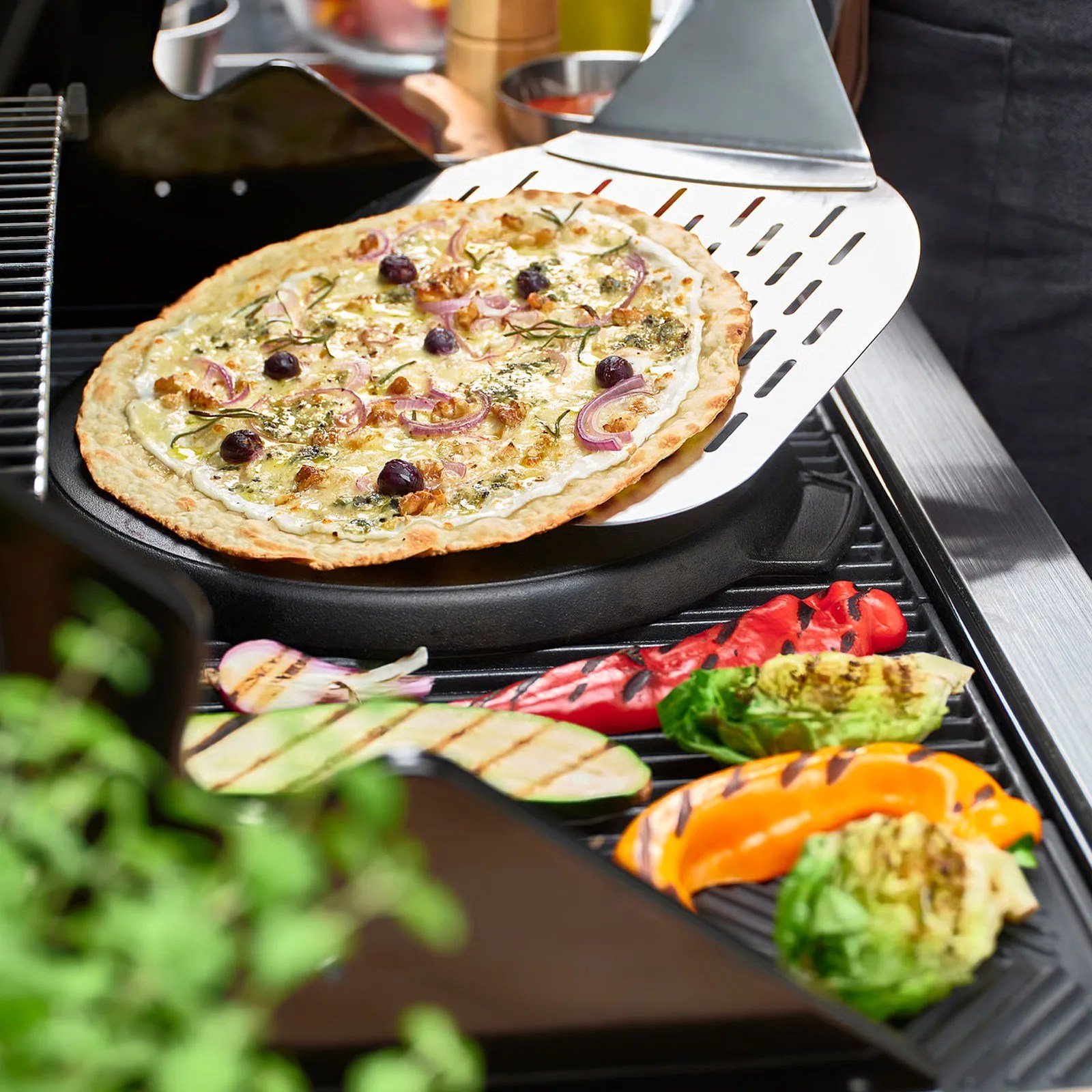 Thin-crust pizza with cheese, red onions, olives, and herbs on a metal pizza peel over a grill with grilled vegetables.