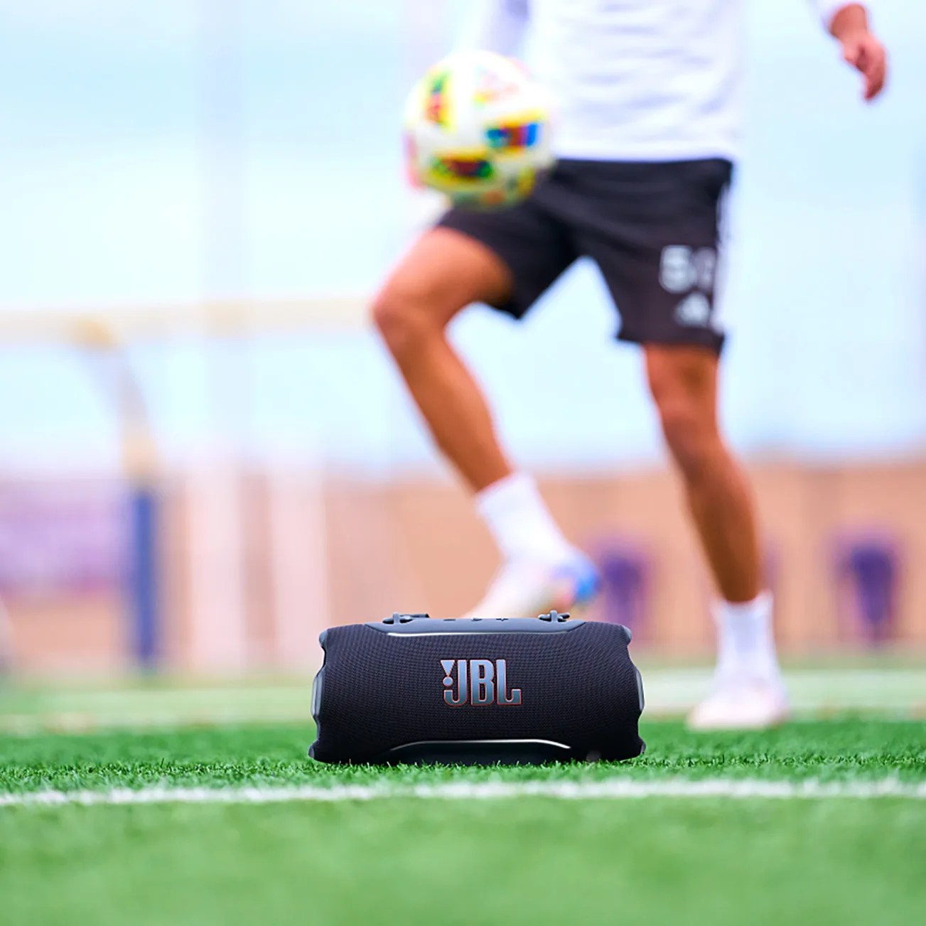Black JBL portable Bluetooth speaker on grass with a person playing soccer in the background.