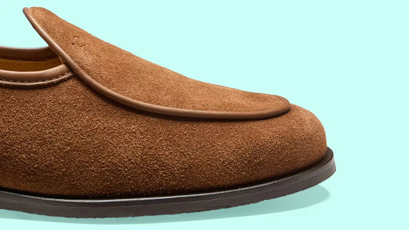 Brown suede loafer with leather trim and black sole on a light blue background.